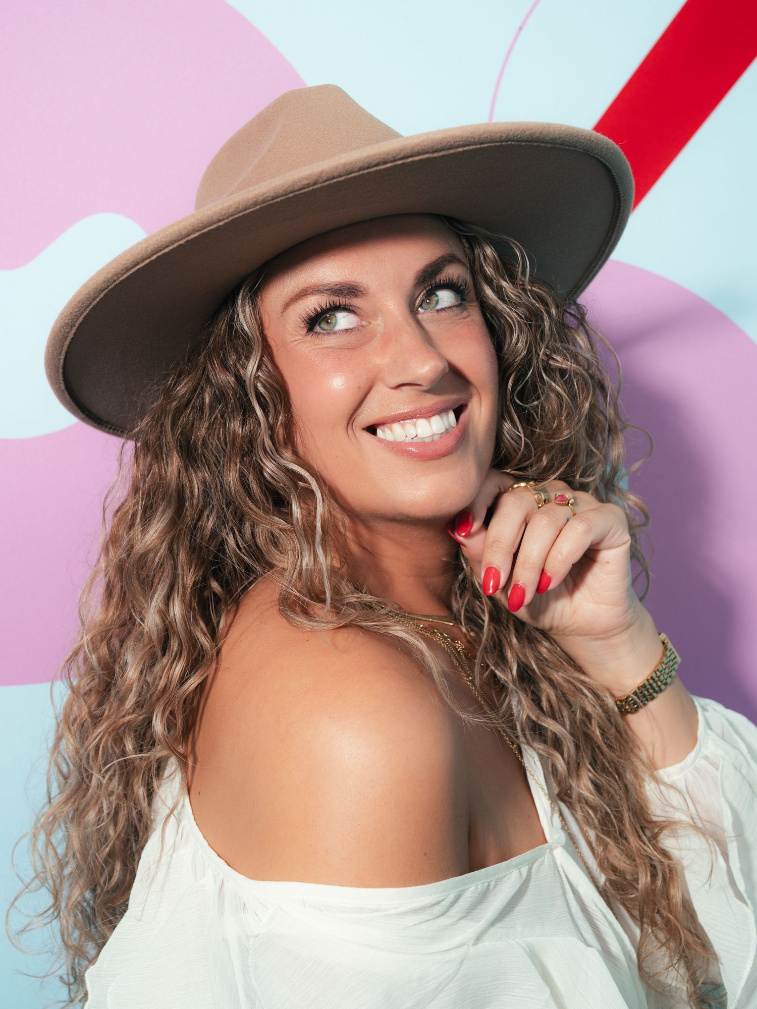 Woman with long curly hair wearing a wide-brim tan hat and white off-shoulder top, smiling against colorful backdrop.