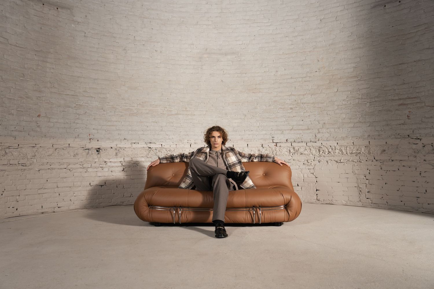 Someone sits in the middle of a large tan leather couch in a minimalist white room with brick walls.