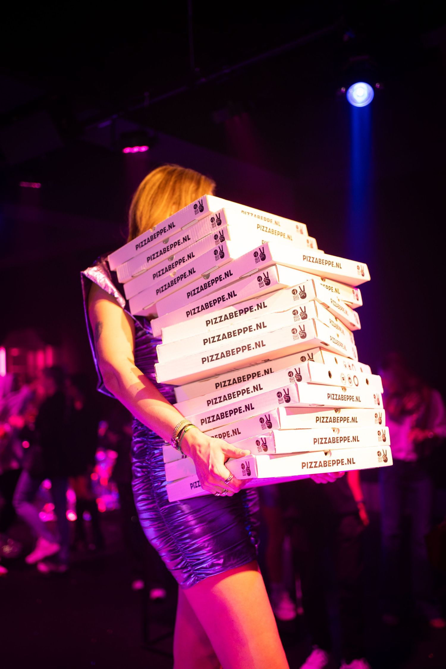 Woman in silver dress carrying a tall stack of Pizzabeppe.nl pizza boxes at a nightclub event.