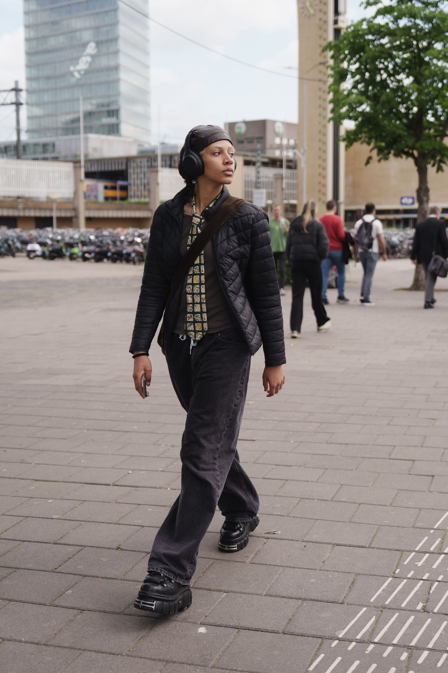 Young woman in black quilted jacket, wide-leg pants, and platform shoes walks through an urban plaza wearing headphones.