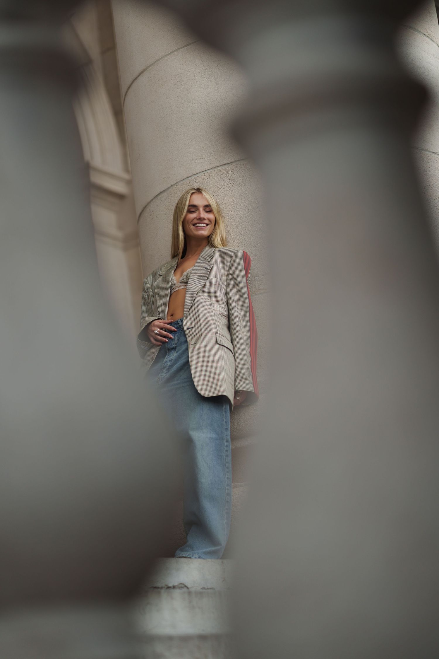 Smiling blonde woman in oversized blazer and wide-leg jeans leaning against stone columns outdoors.