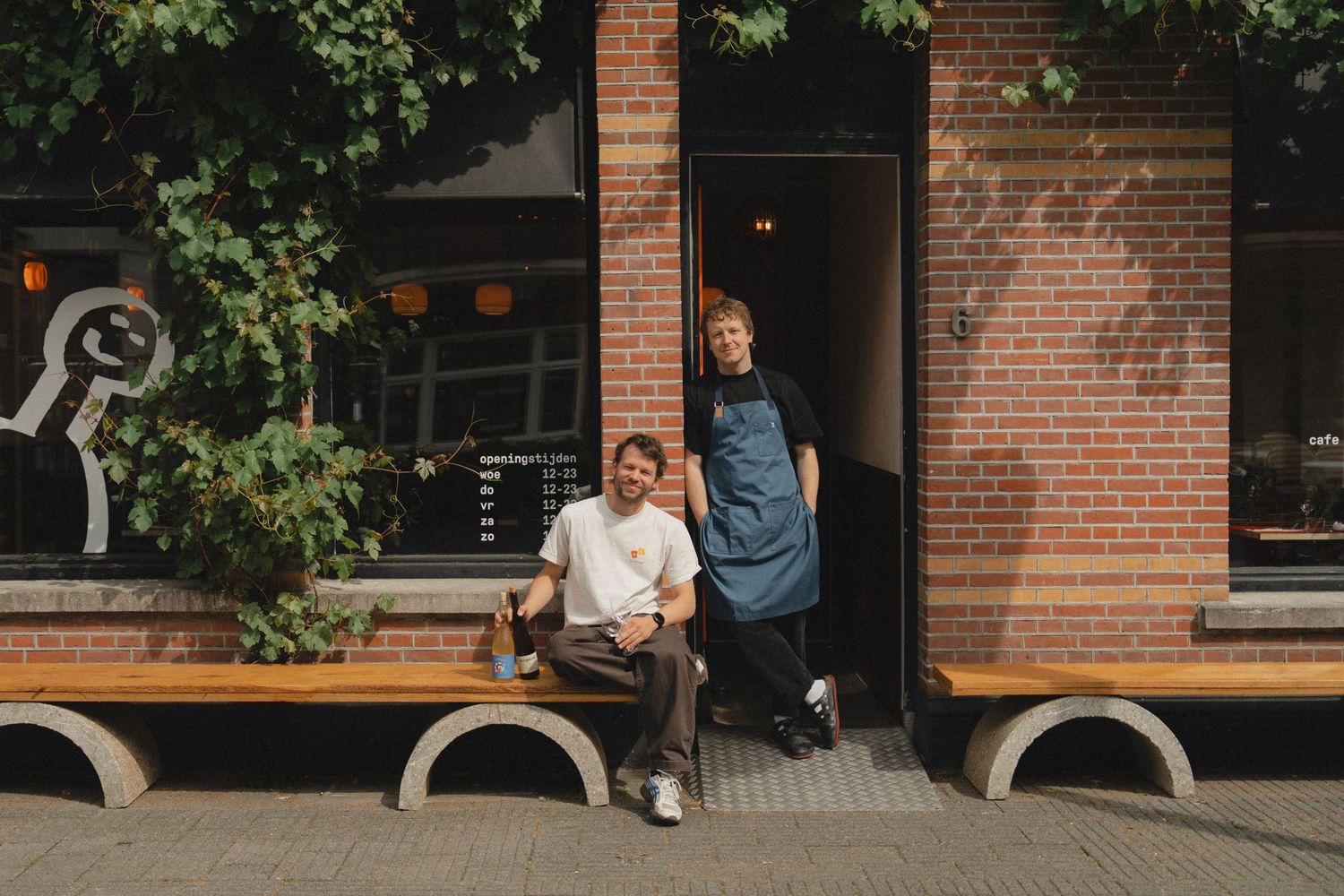Two people pose outside a brick building with lush greenery, one seated on a bench, the other standing in a blue apron.