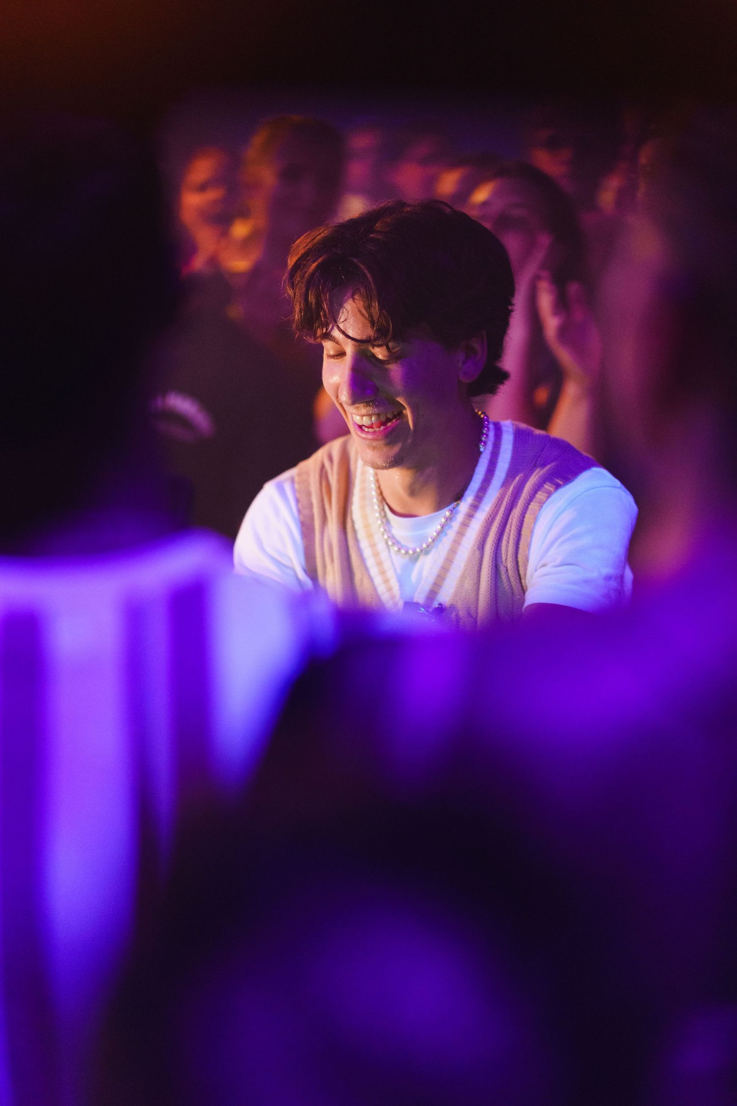 A young man smiling under purple stage lights surrounded by an energetic concert crowd.