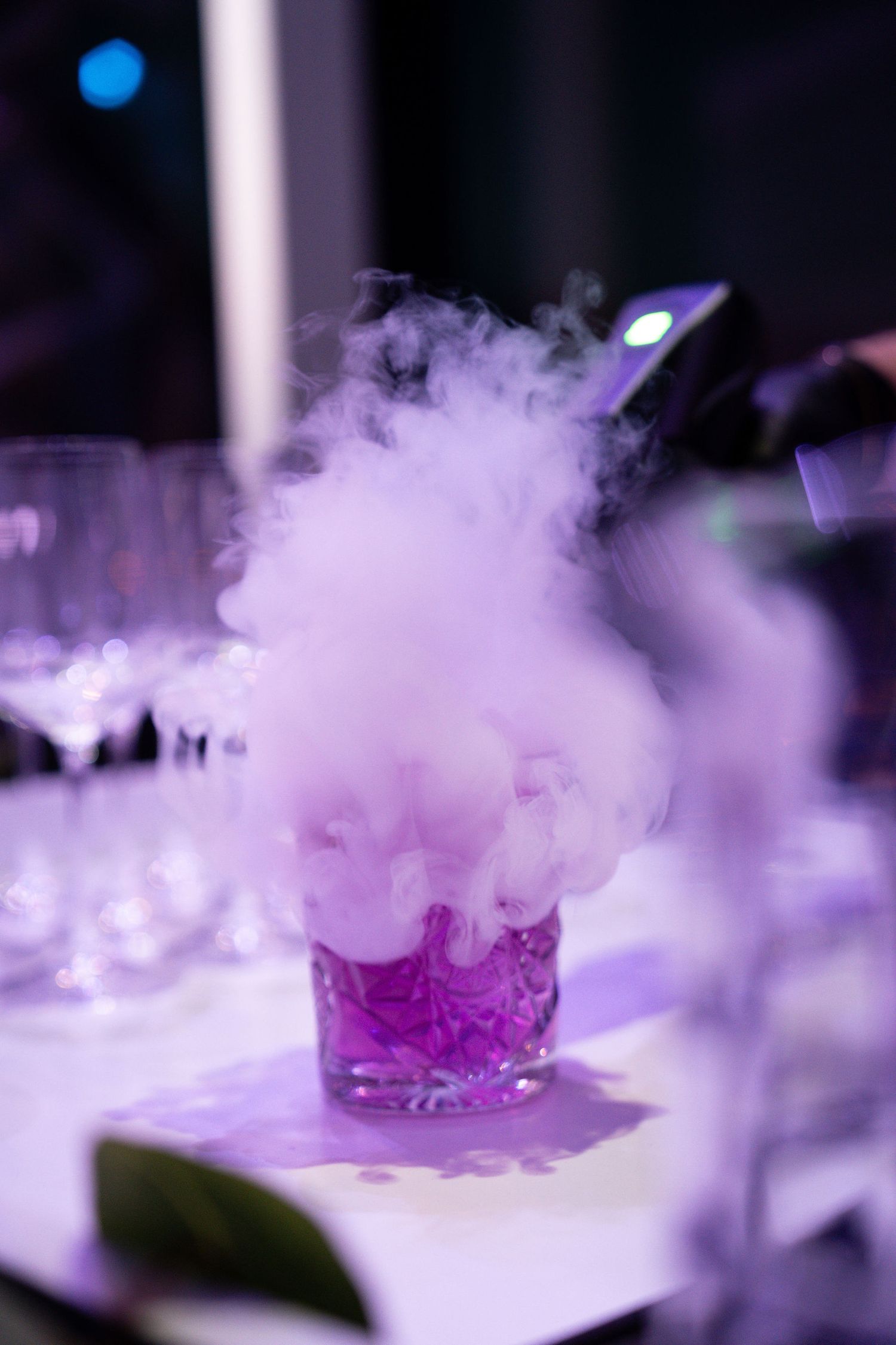 A dramatic purple cocktail billowing with dry ice smoke sits on a white surface at an elegant bar or event.