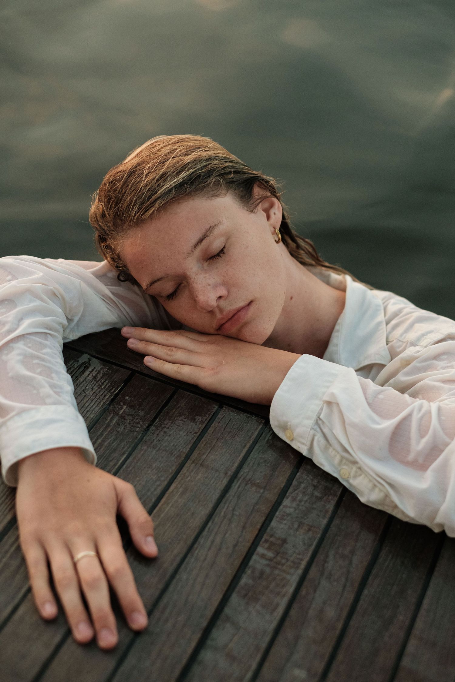 Woman in wet white shirt resting head on wooden dock by water, eyes closed, wearing a ring.