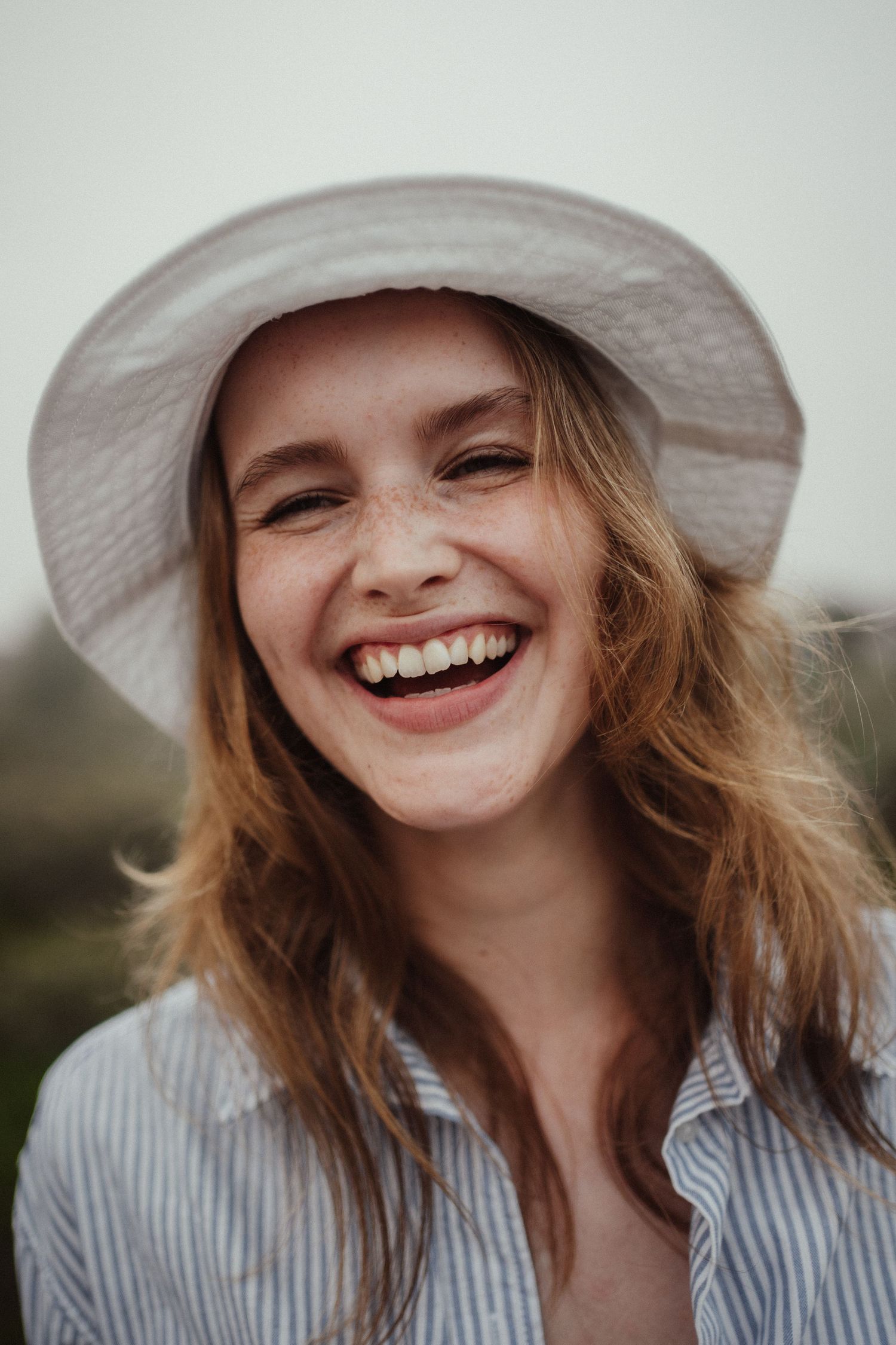 Laughing young woman with freckles wearing a white sun hat and striped shirt outdoors.