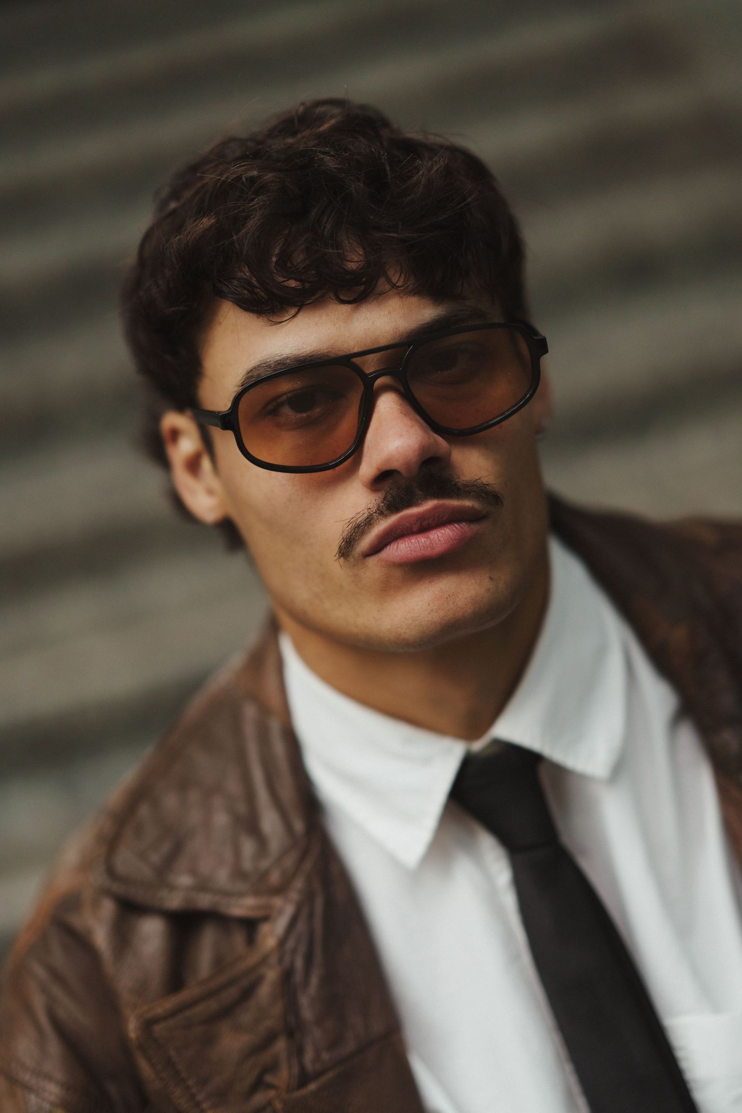 Man with mustache wearing brown leather jacket, white shirt, black tie, and retro tinted sunglasses outdoors.
