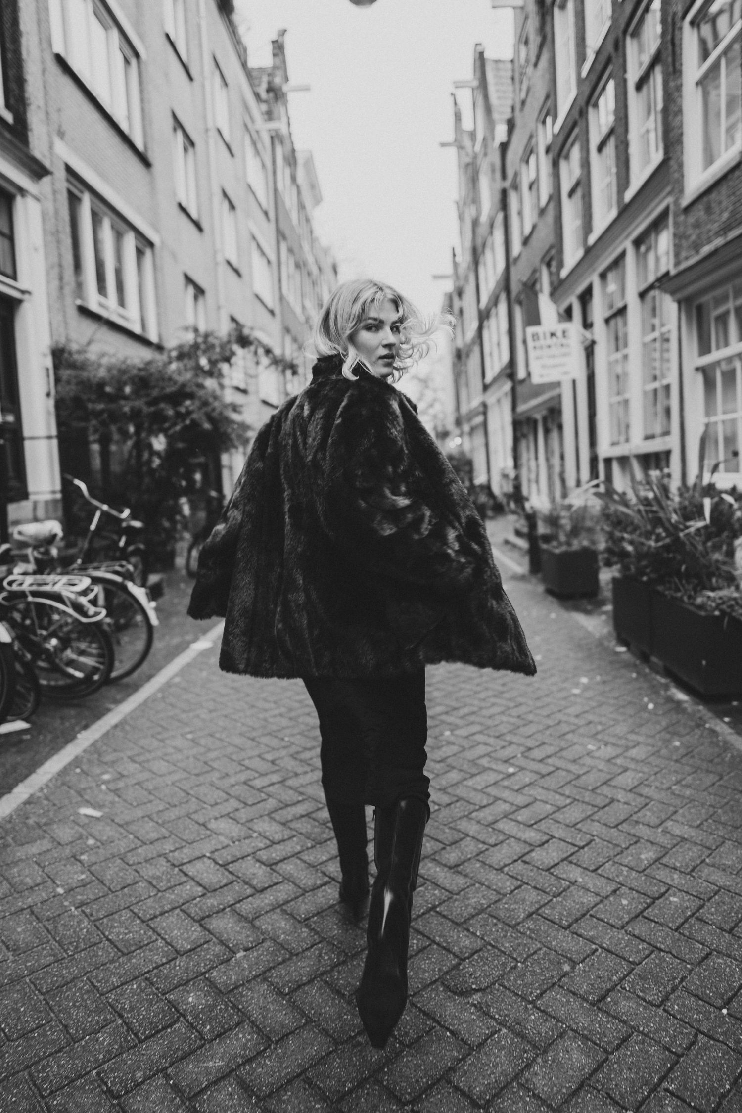 Blonde woman in black fur coat walks cobblestone Amsterdam street in black and white photo.