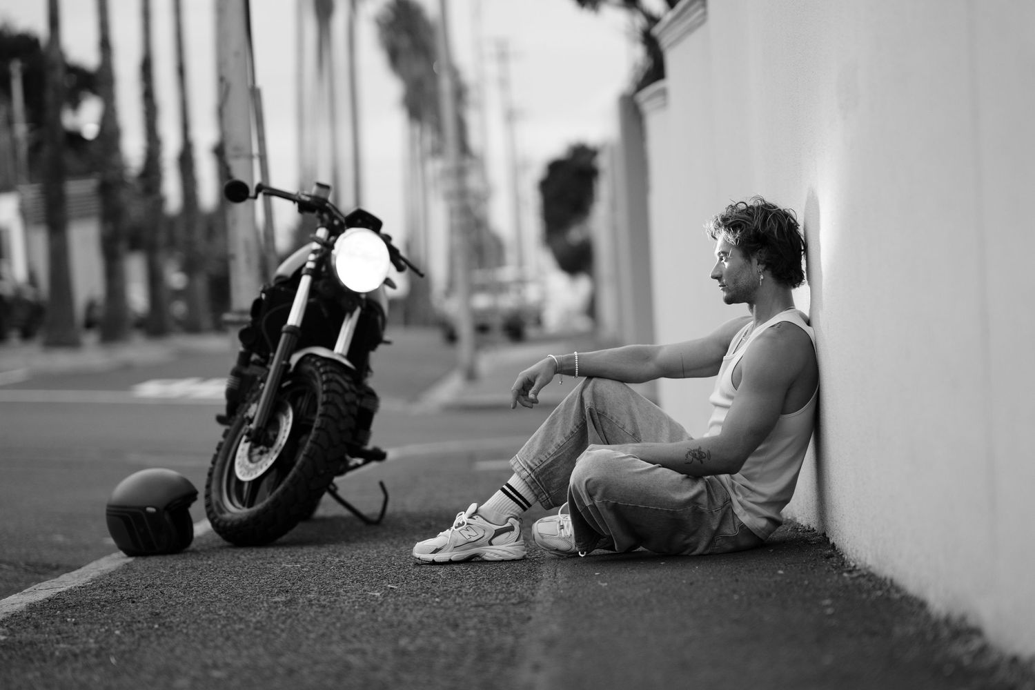 Black and white photo of a man sitting against a wall next to a motorcycle on a city street.