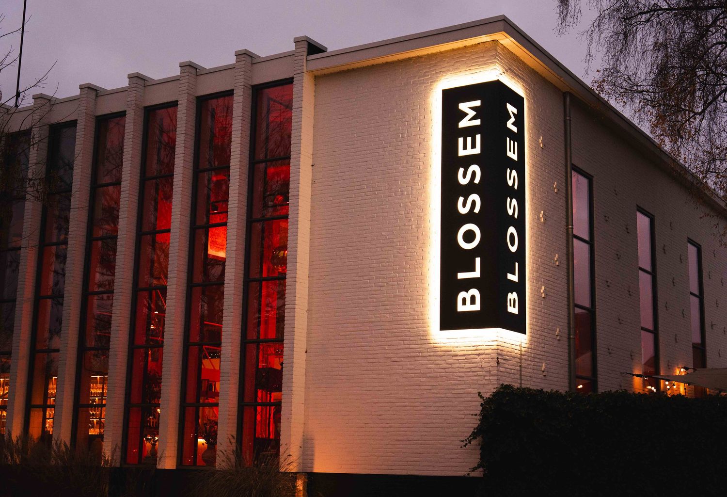 Blossom restaurant exterior illuminated at dusk with glowing red windows and vertical signage on white building.