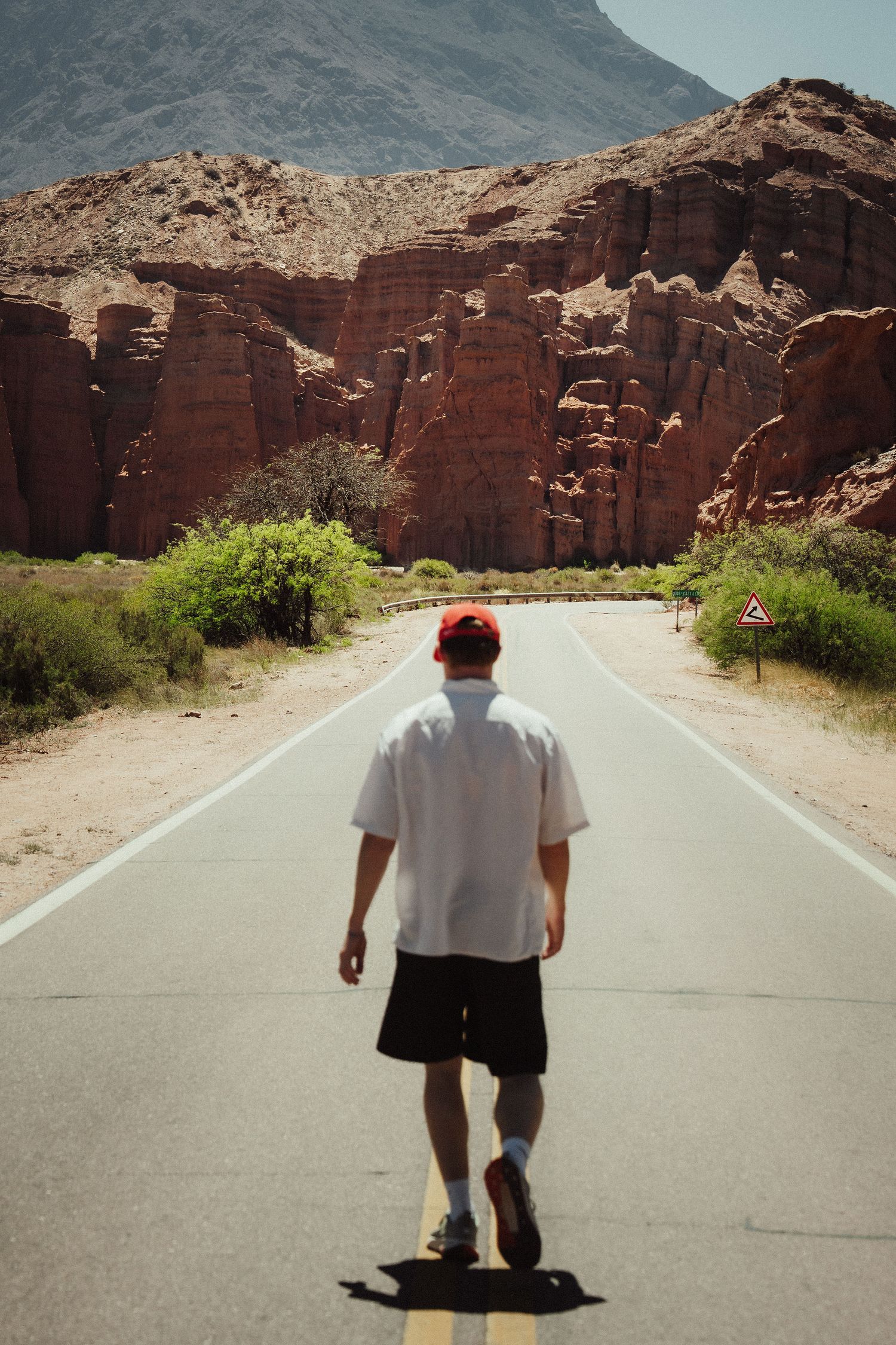 Man in white shirt and red cap walks down desert road toward dramatic red rock canyon formations.