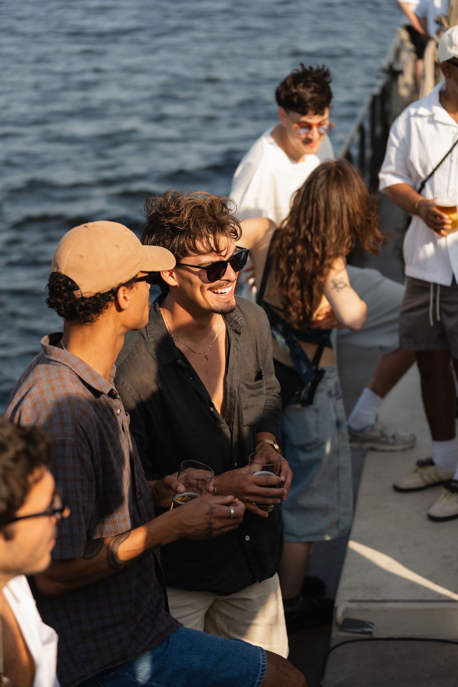 A group of young people socializing and laughing on a boat deck at sunset with sparkling water in the background.