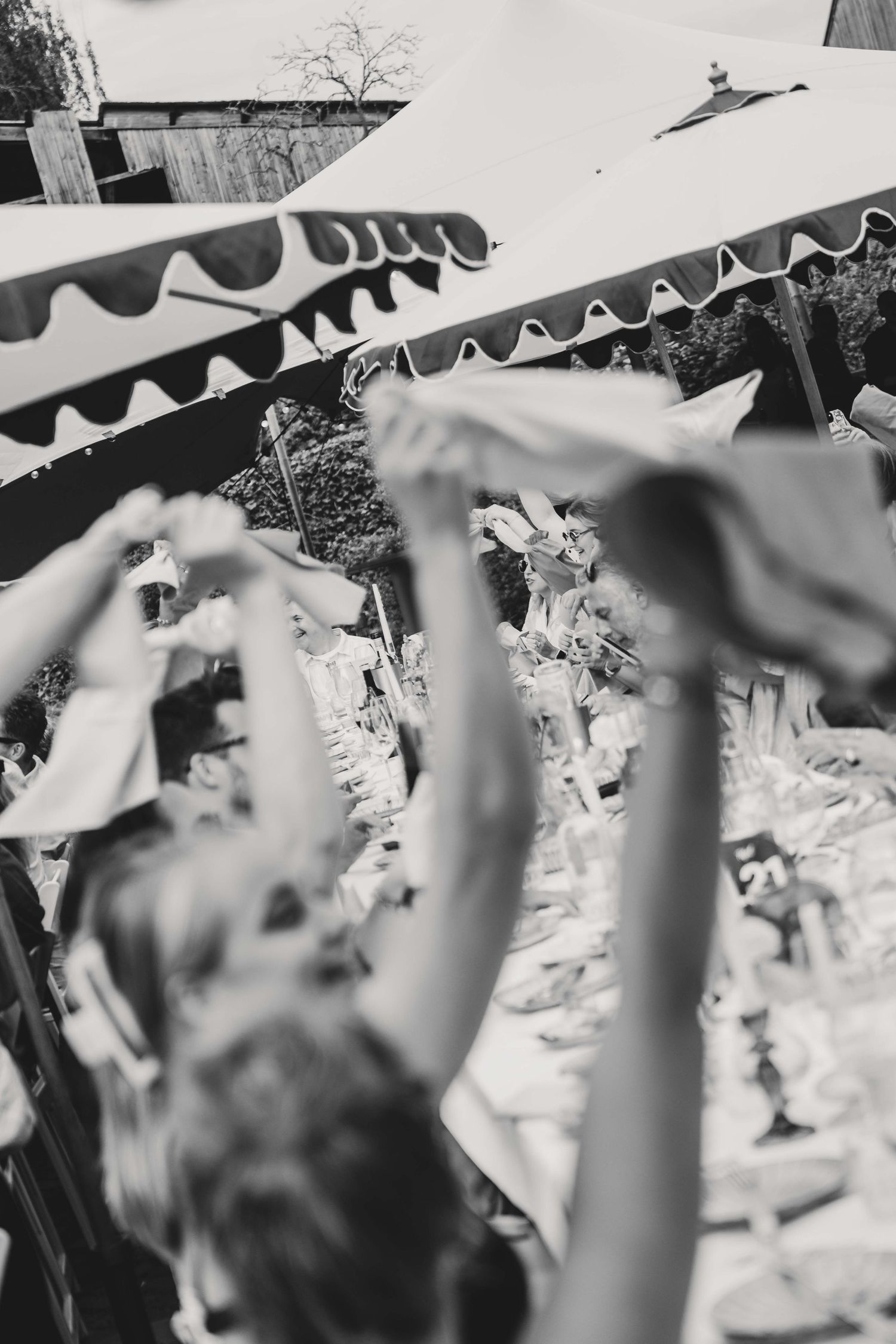 Black and white photo of guests waving napkins at an outdoor wedding reception under striped tent.