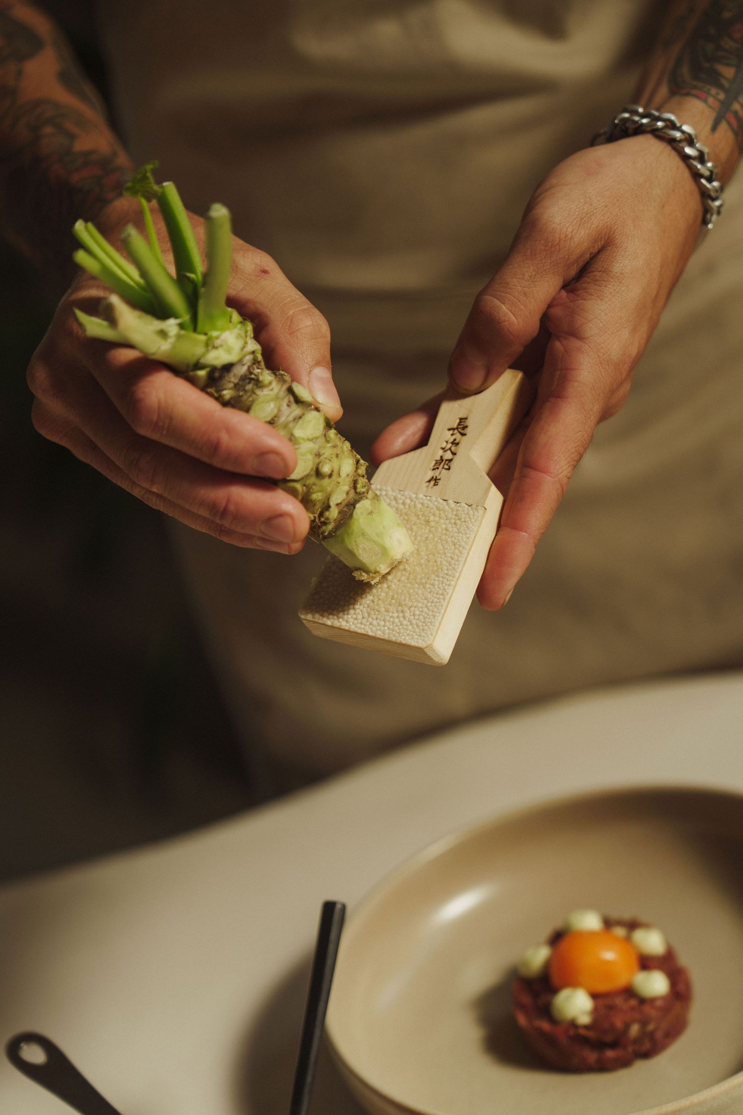 Tattooed chef grating fresh wasabi root over a plate with beef tartare and egg yolk in a fine dining restaurant.