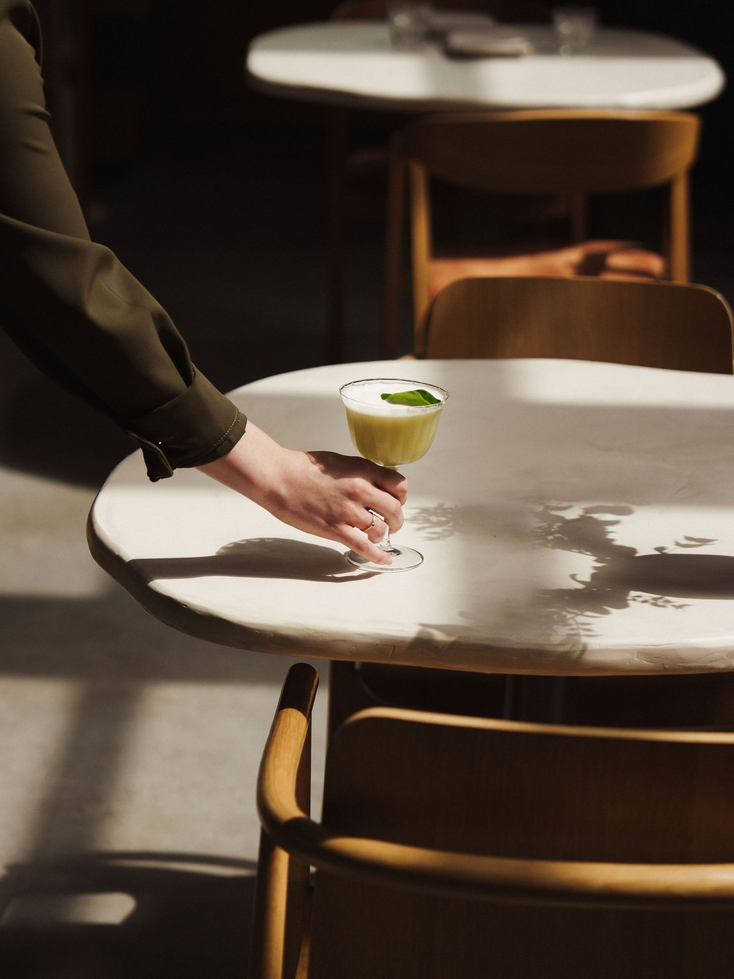 A hand places a green cocktail in a coupe glass on a white marble table in a moody, sunlit cafe setting.