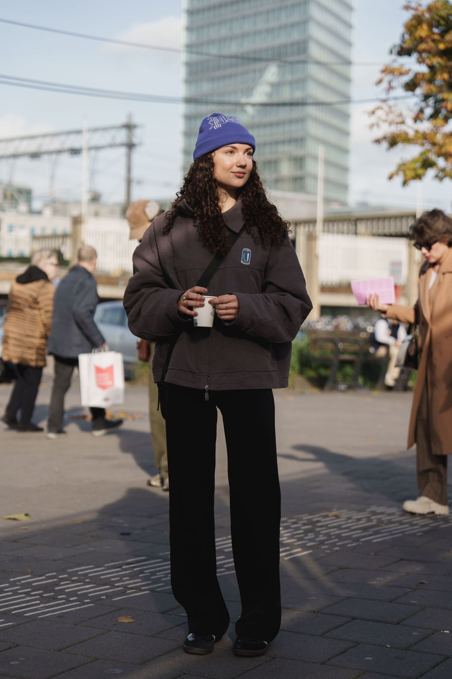 Woman in blue beanie, dark puffer jacket, and black wide-leg pants holding a cup outdoors in an urban setting.