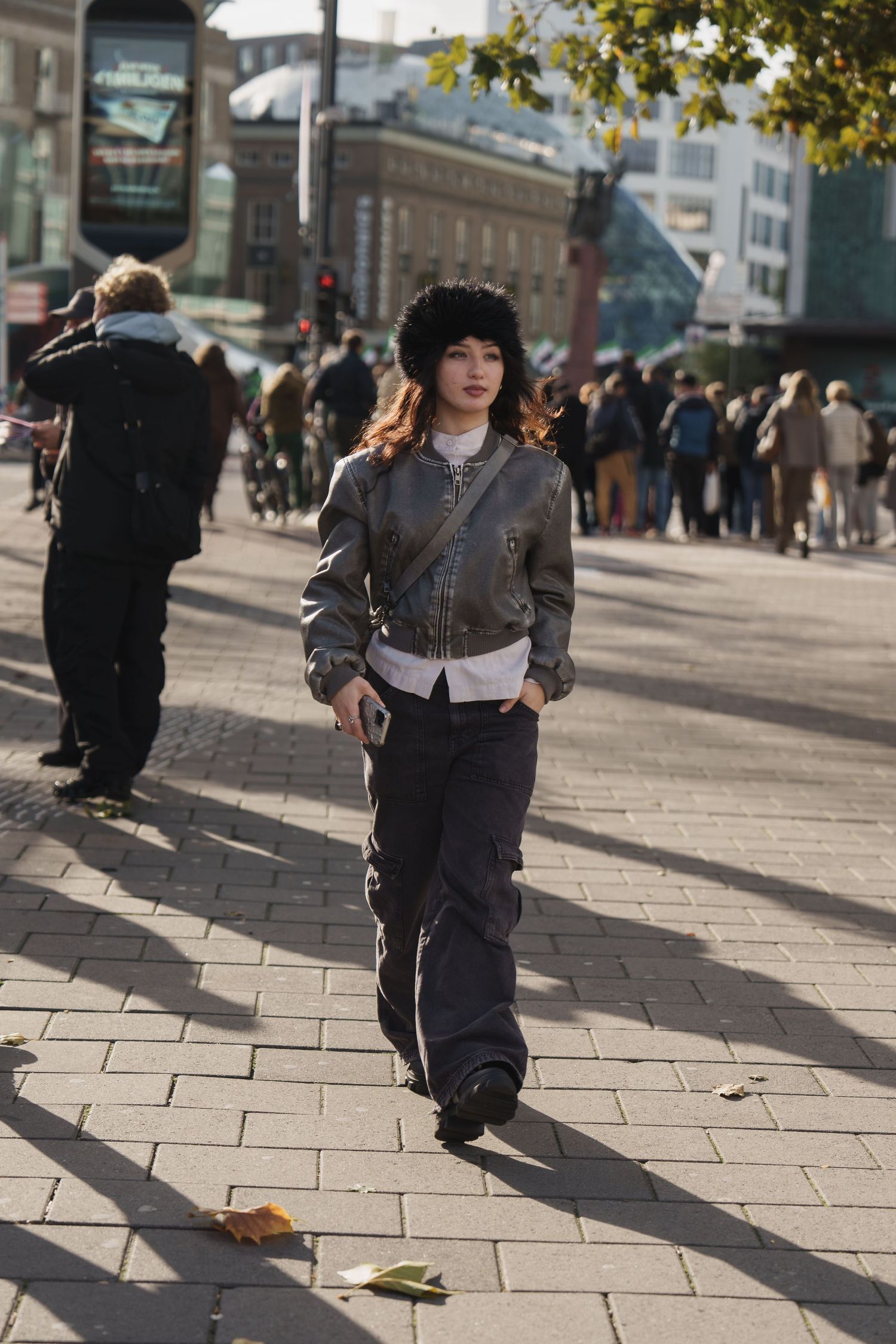 Young woman in bomber jacket and fur hat walking on busy city street.