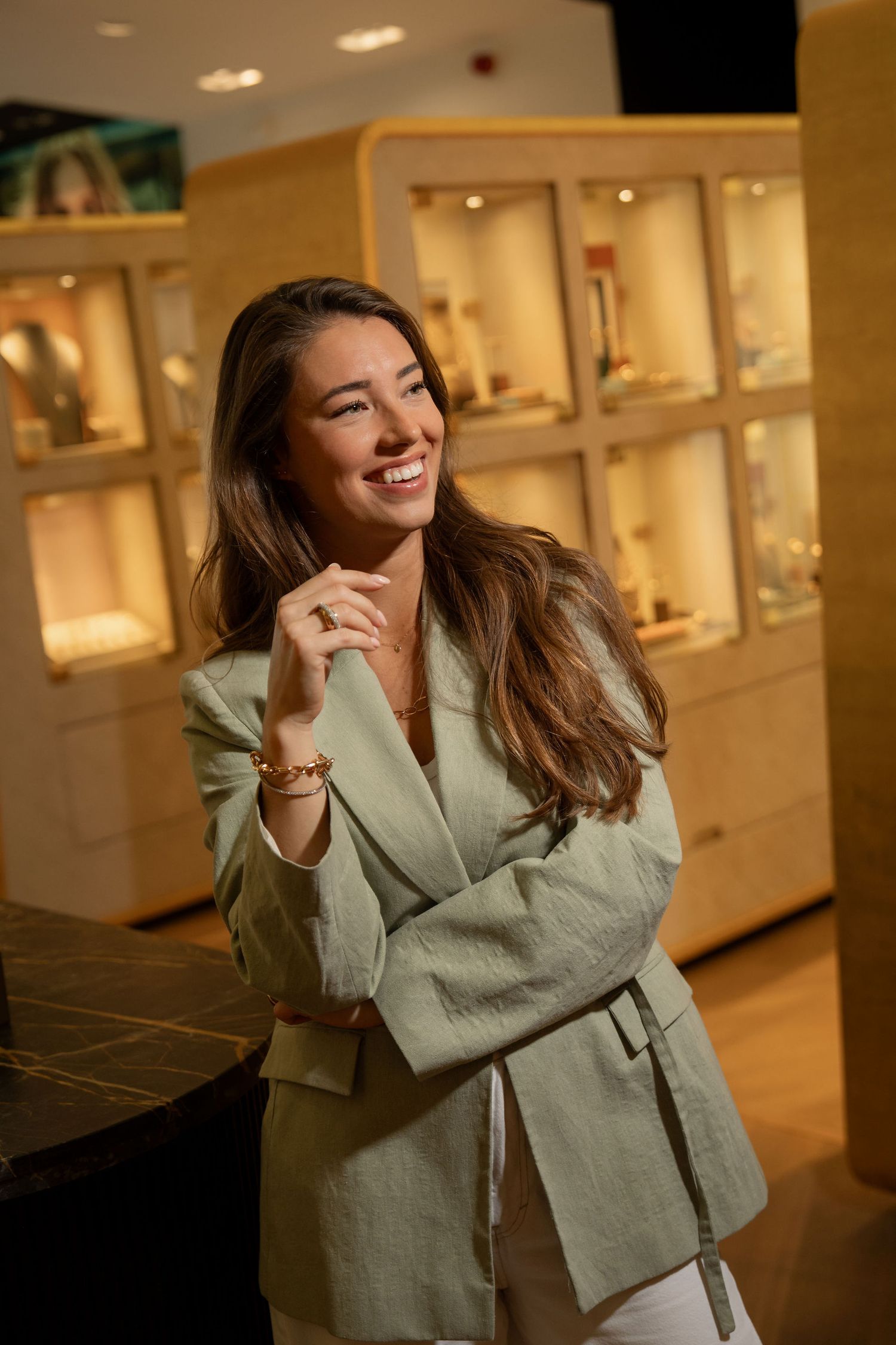Smiling woman in sage green blazer poses in upscale jewelry store with illuminated display cases in background.