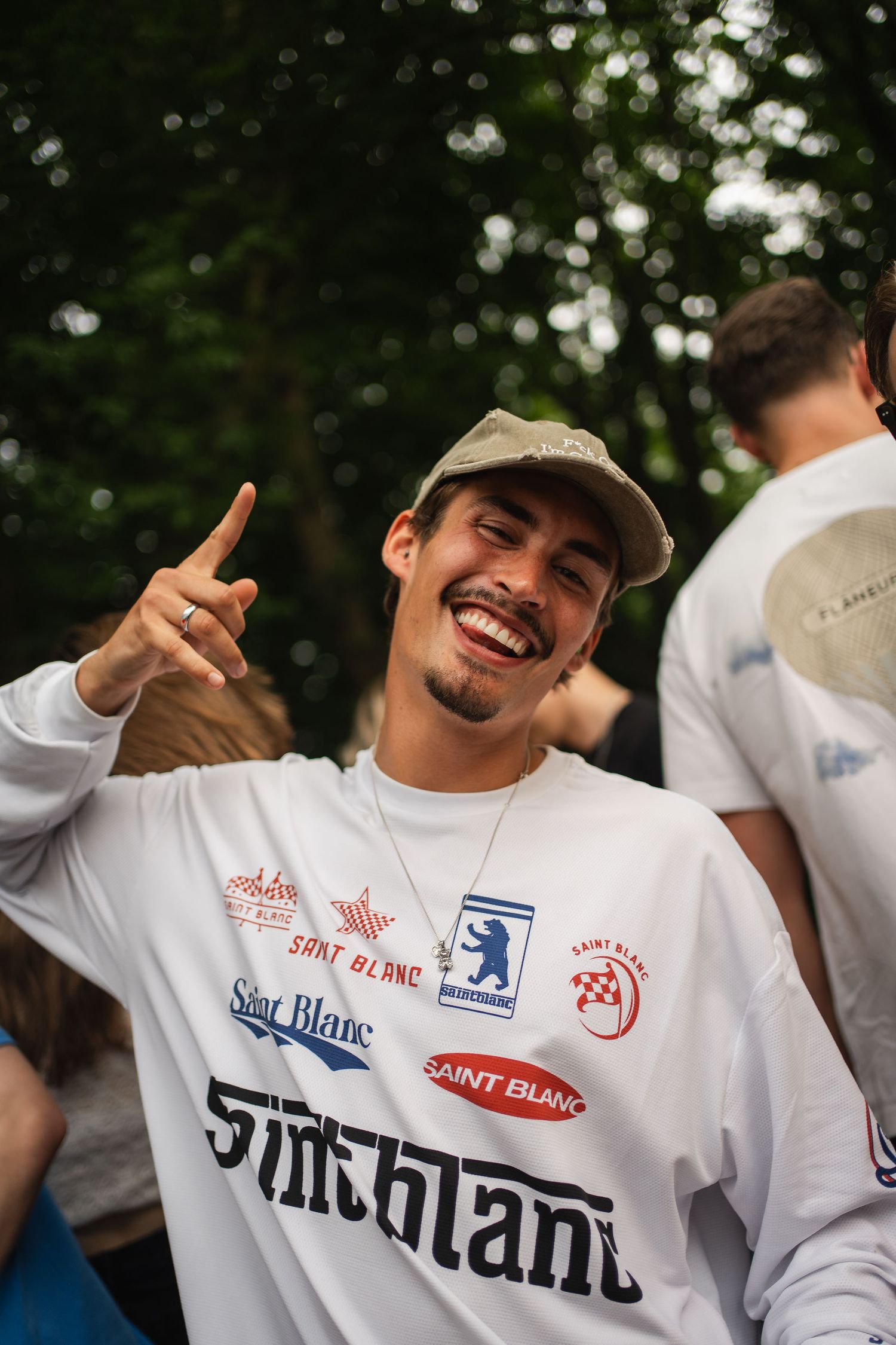 Smiling young man wearing a white Saint Blanc graphic long sleeve shirt and cap, pointing finger gun outdoors.