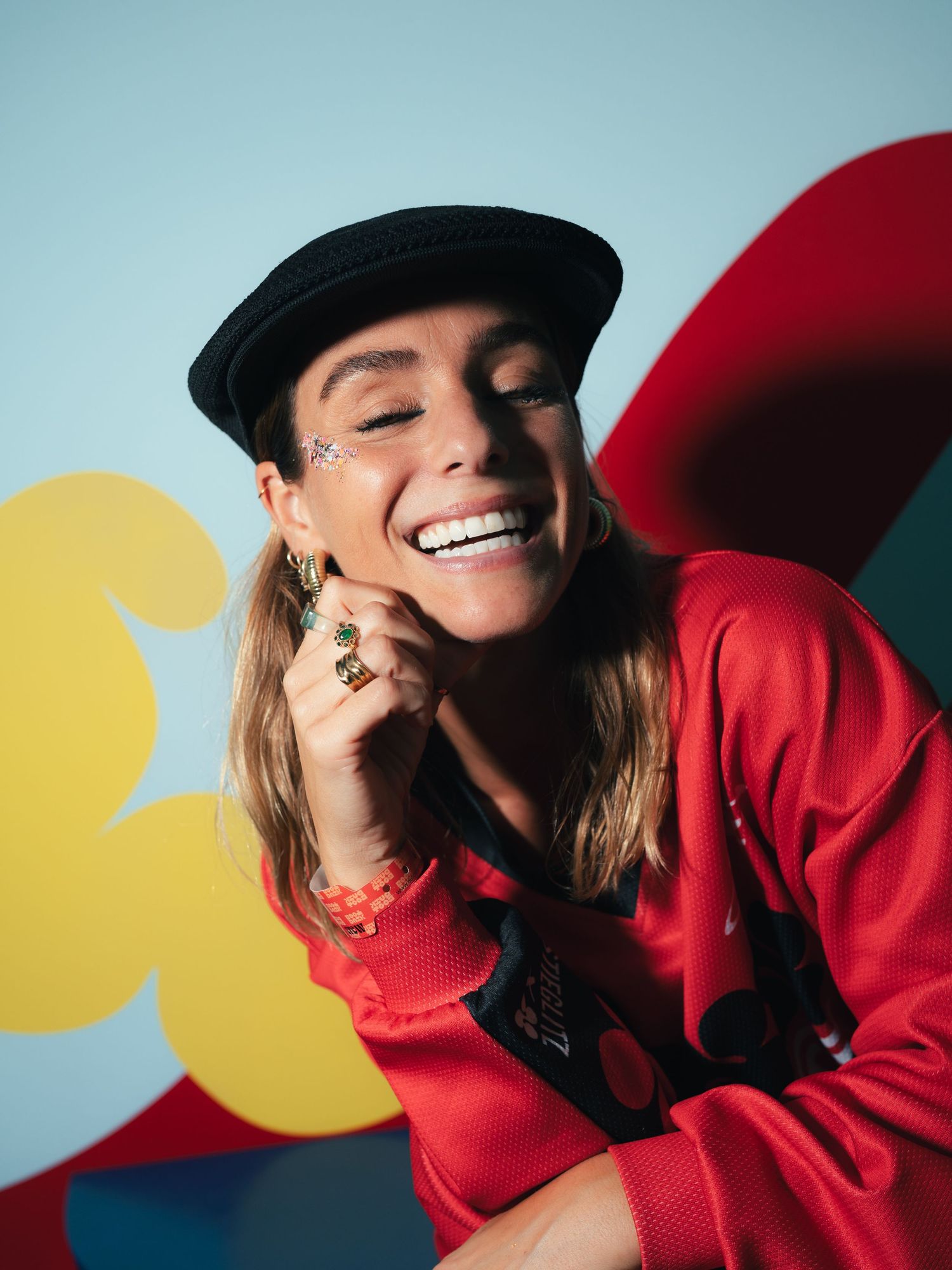 Woman in red jacket and black beret laughing with eyes closed, wearing green rings against colorful backdrop.