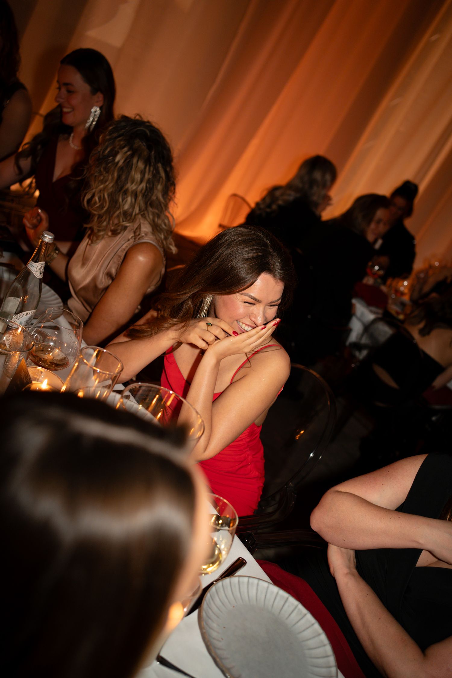 A woman in a red dress laughs joyfully at an elegant evening gala event with guests seated at tables.