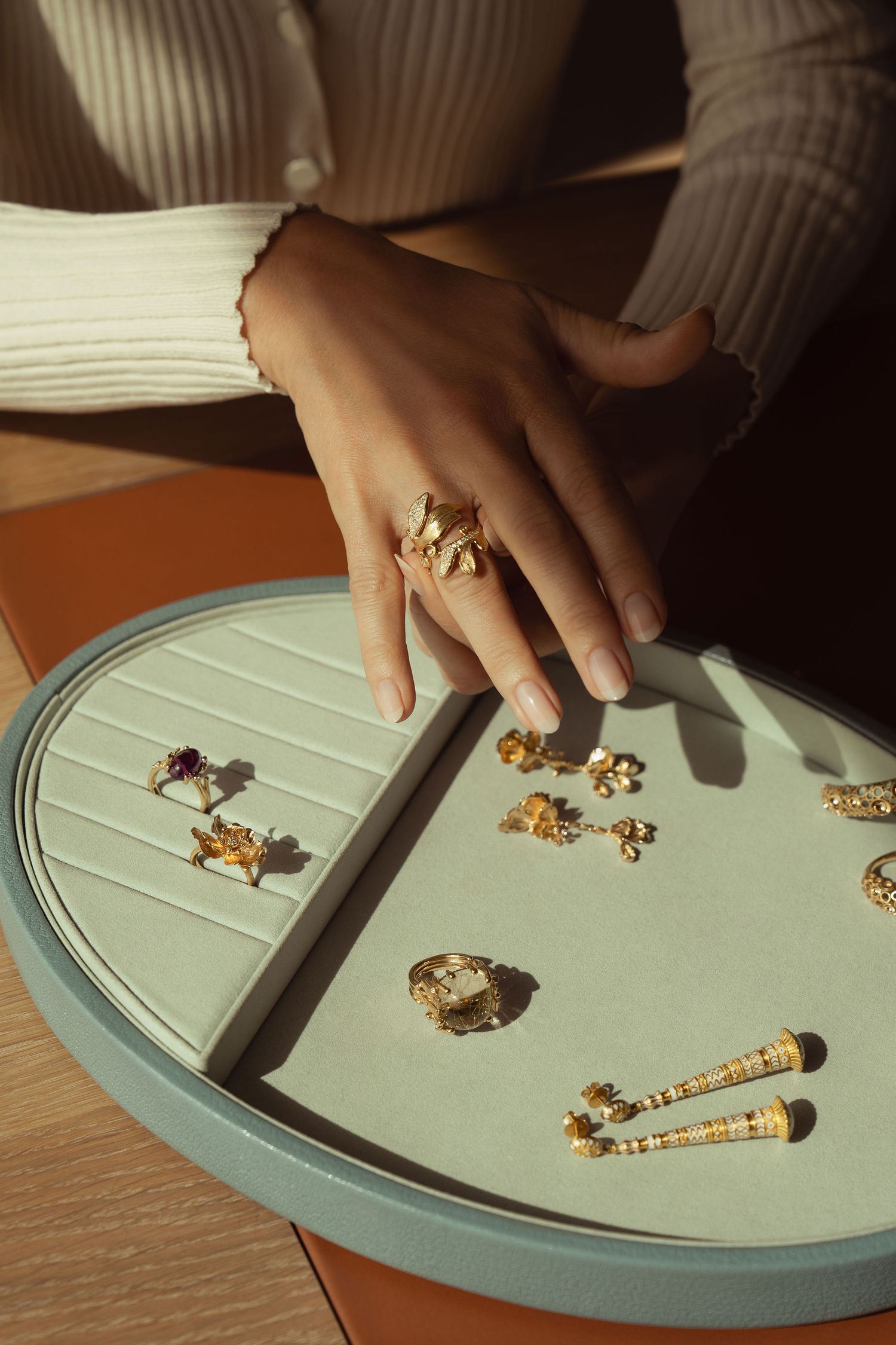 Woman wearing a large gold statement ring displays luxury gold jewelry pieces on a mint green velvet tray in warm sunlight.