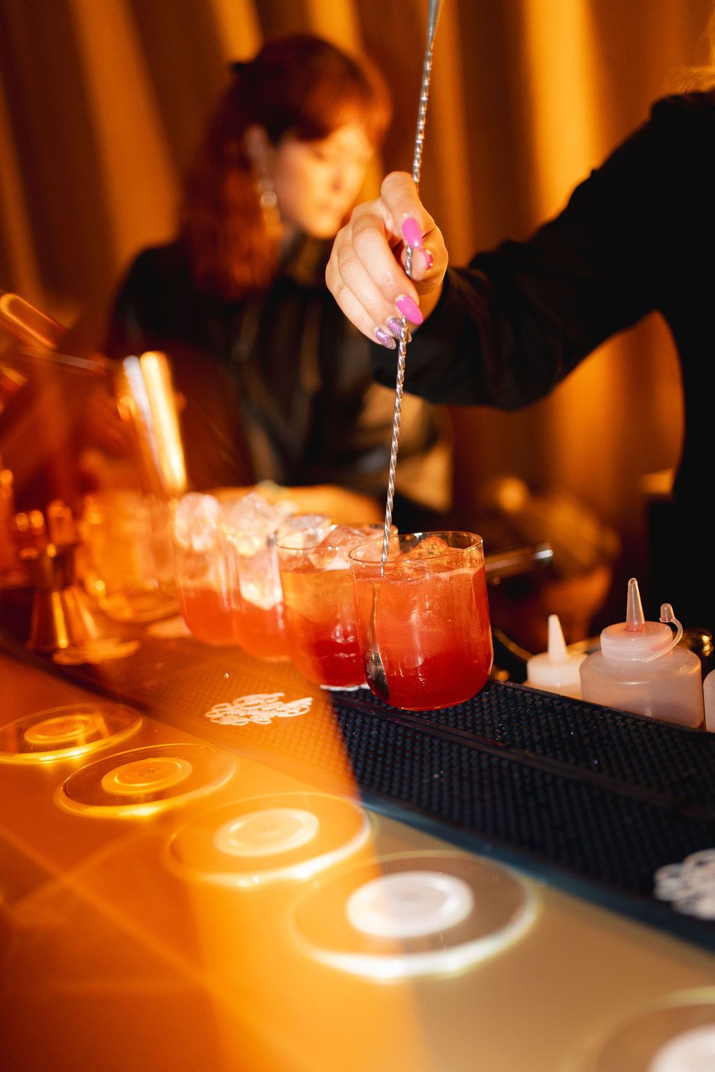 Bartender pouring red cocktails into glasses on a bar counter with warm orange lighting at a nightclub event.
