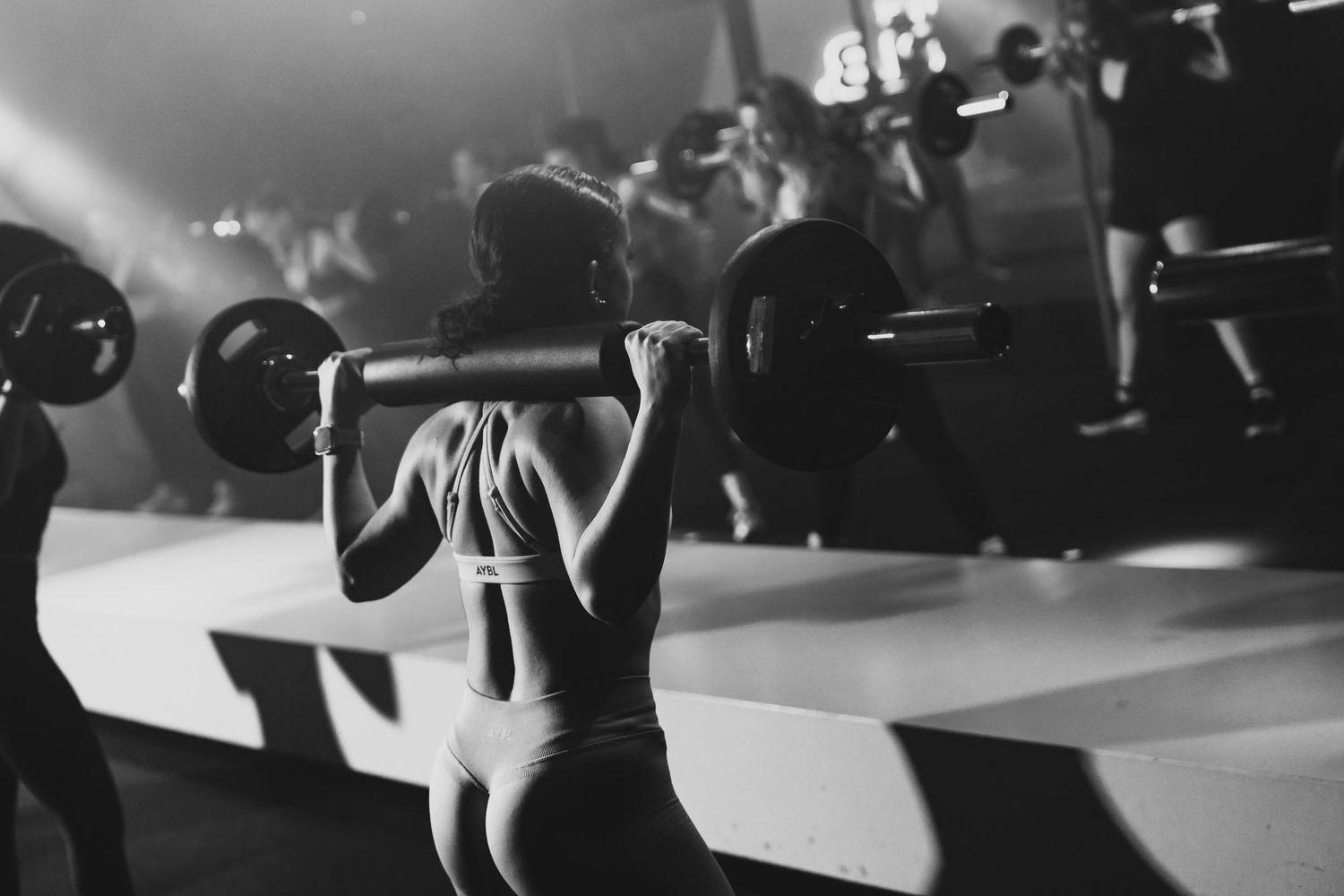 Woman performing barbell squats in a busy gym, black and white fitness photography.