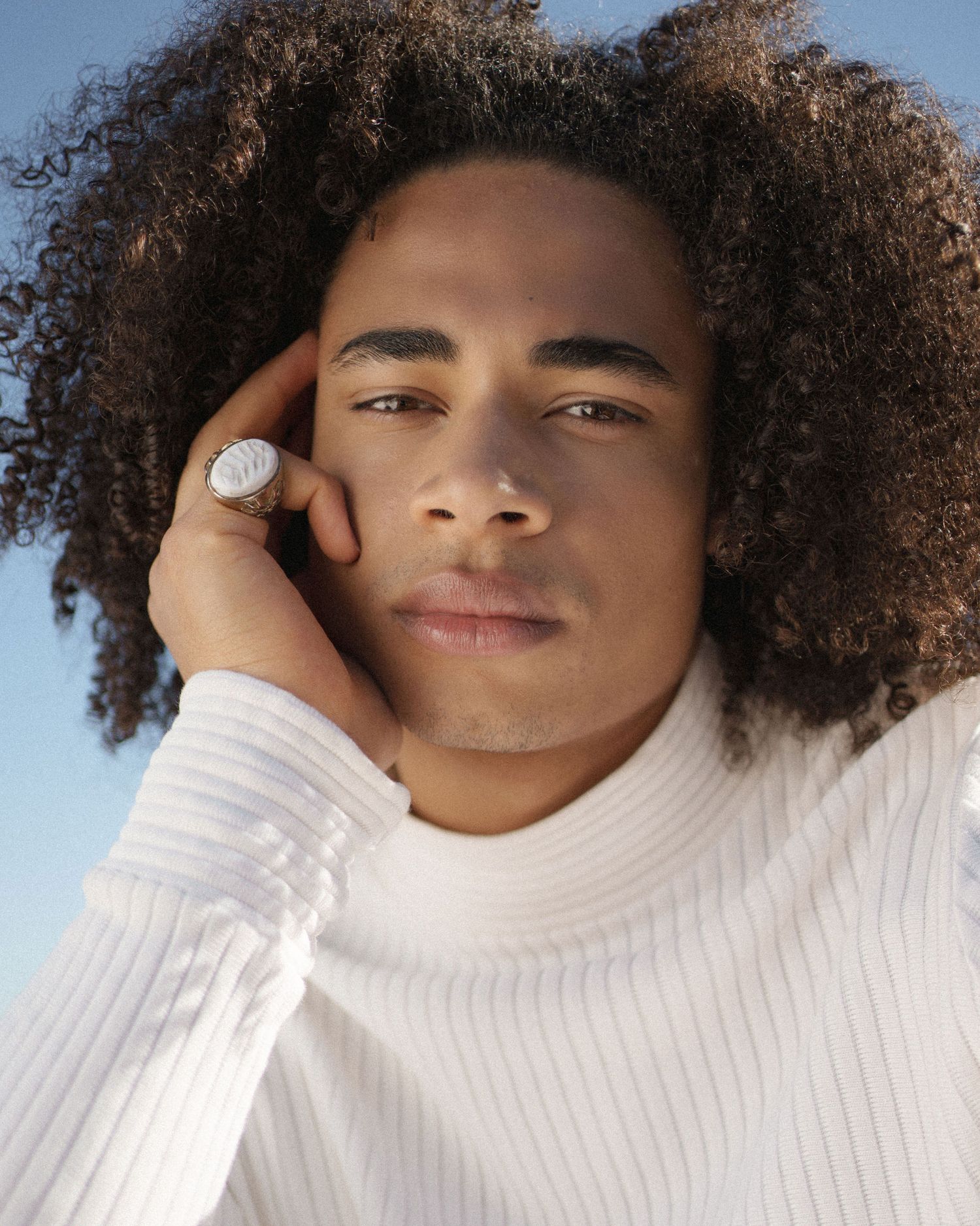 Young man with curly hair wearing a white ribbed turtleneck sweater and silver ring, posing against a blue sky.