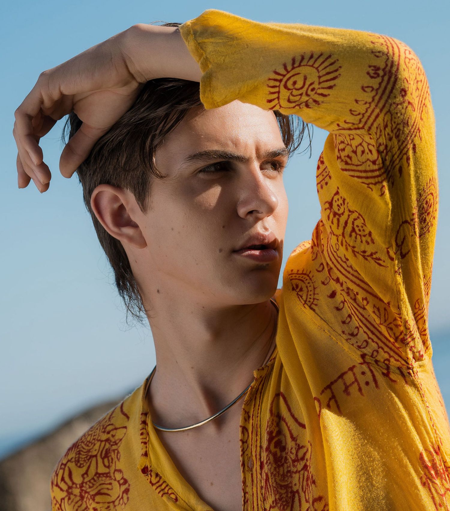 Young man in yellow patterned robe with hand raised to forehead posing outdoors against blue sky.