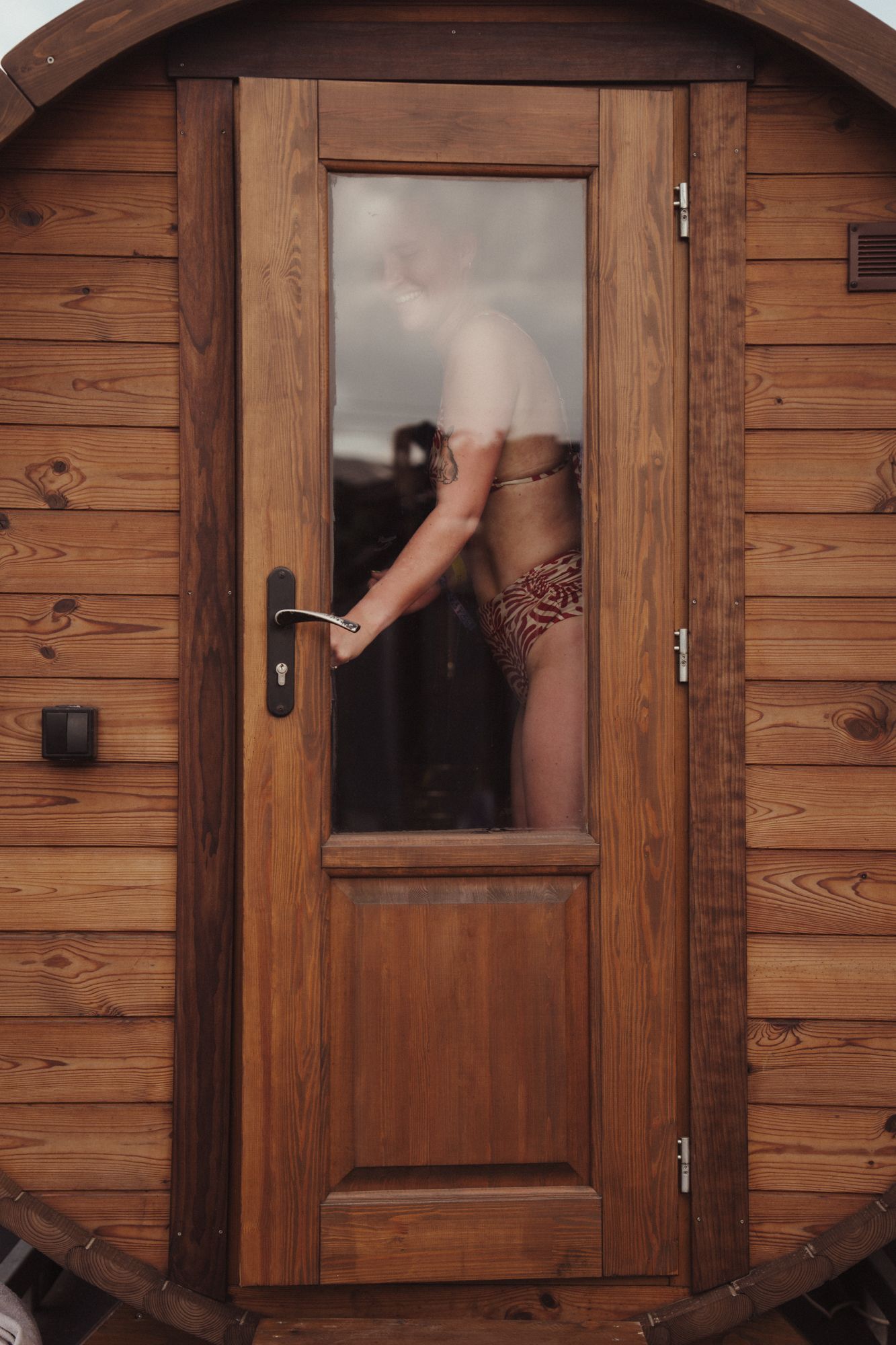 Person in swimwear opening the glass door of a wooden barrel sauna cabin.