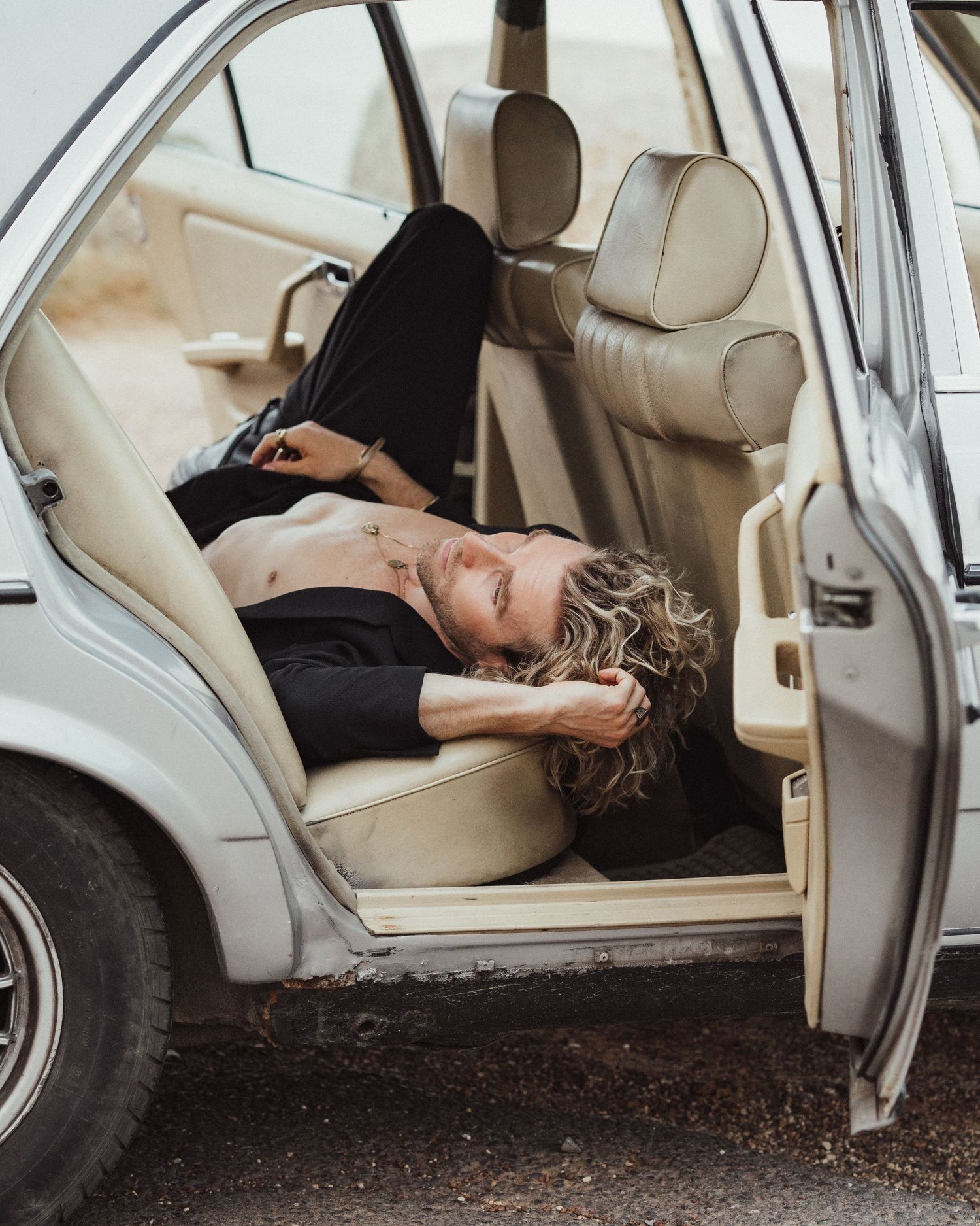 Man with curly hair lying across cream leather backseat of a classic vintage car.