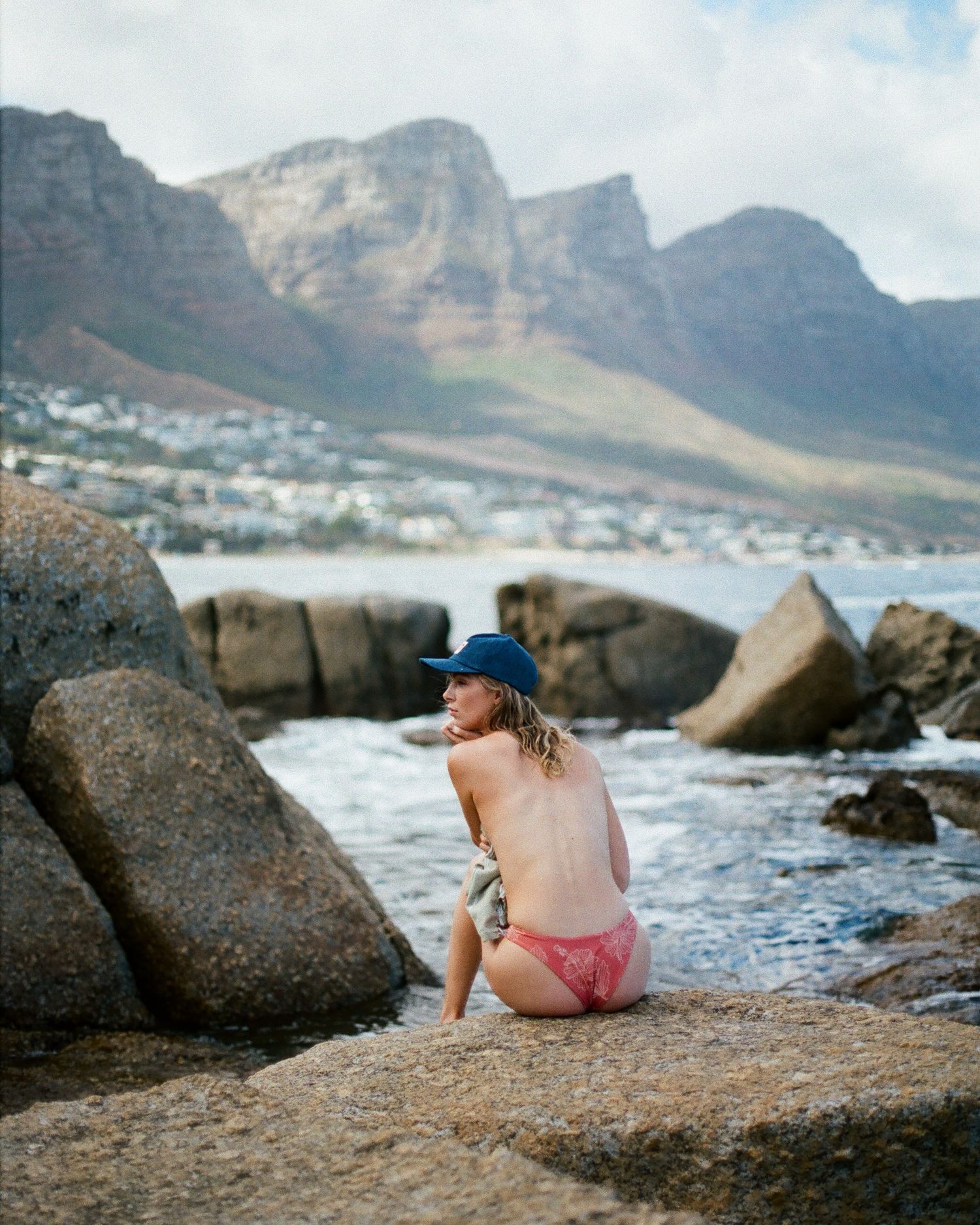 Woman in blue hat and pink bikini sitting on rocks by the ocean with mountain backdrop in Cape Town.