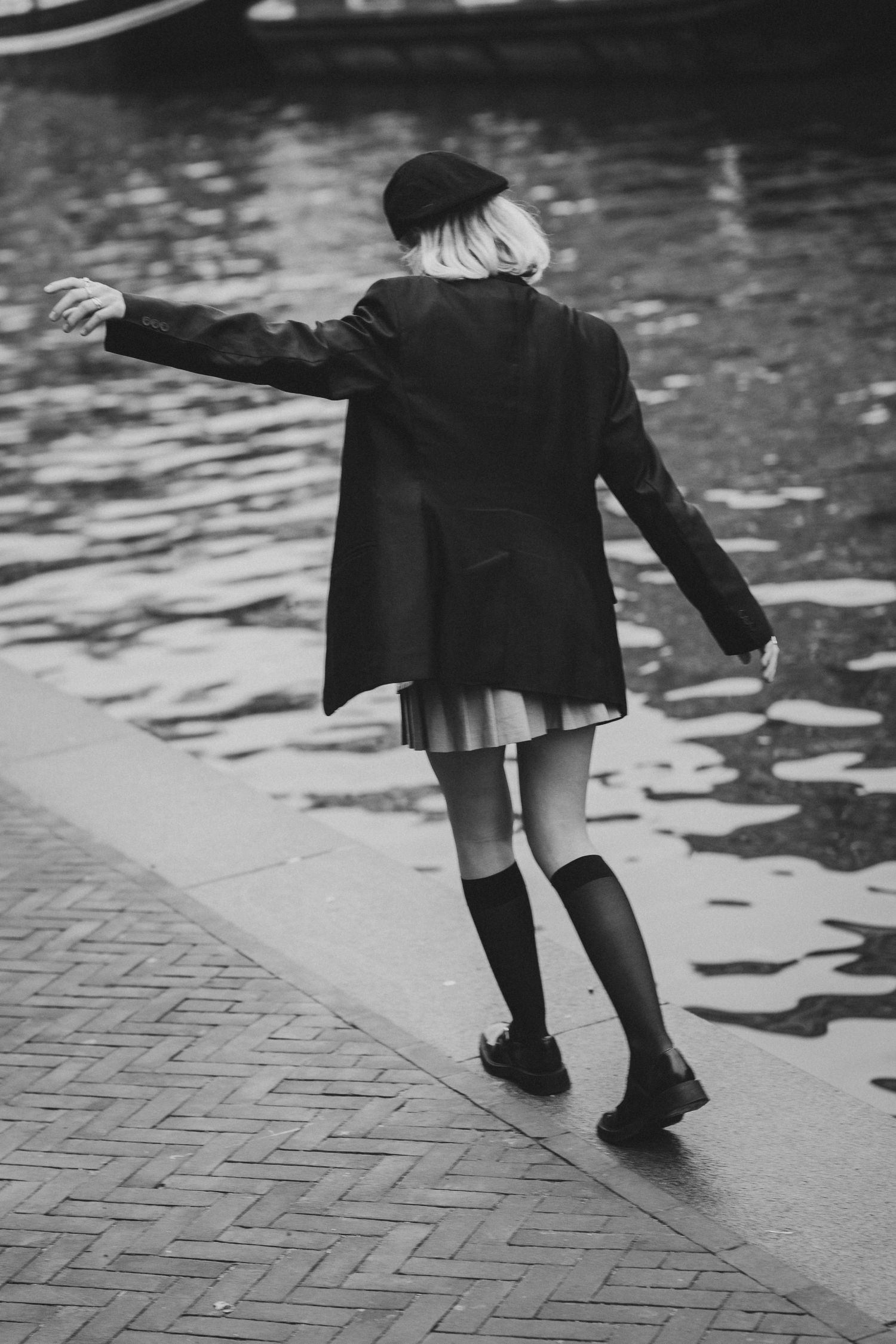 Black and white photo of a woman in beret, blazer, mini skirt and knee socks balancing on canal edge.
