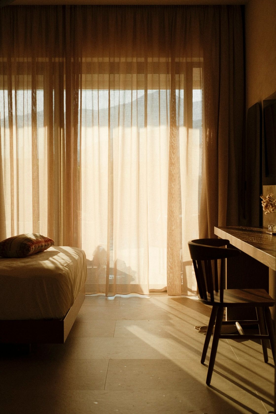 Sunlit hotel room with sheer curtains, a single bed with pillows, wooden chair, and desk bathed in warm golden light.