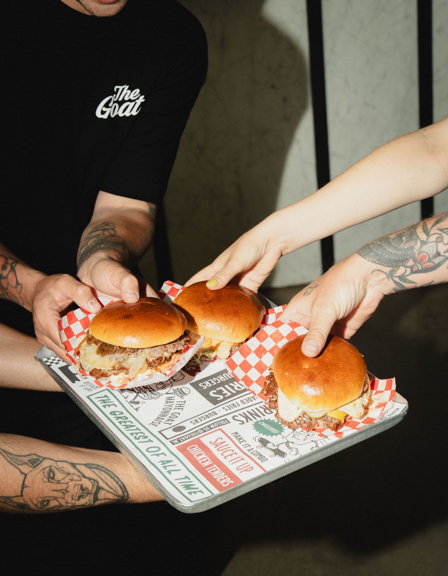 Tattooed hands holding a tray of three gourmet burgers on checkered paper, with a person wearing a The Goat black tee.