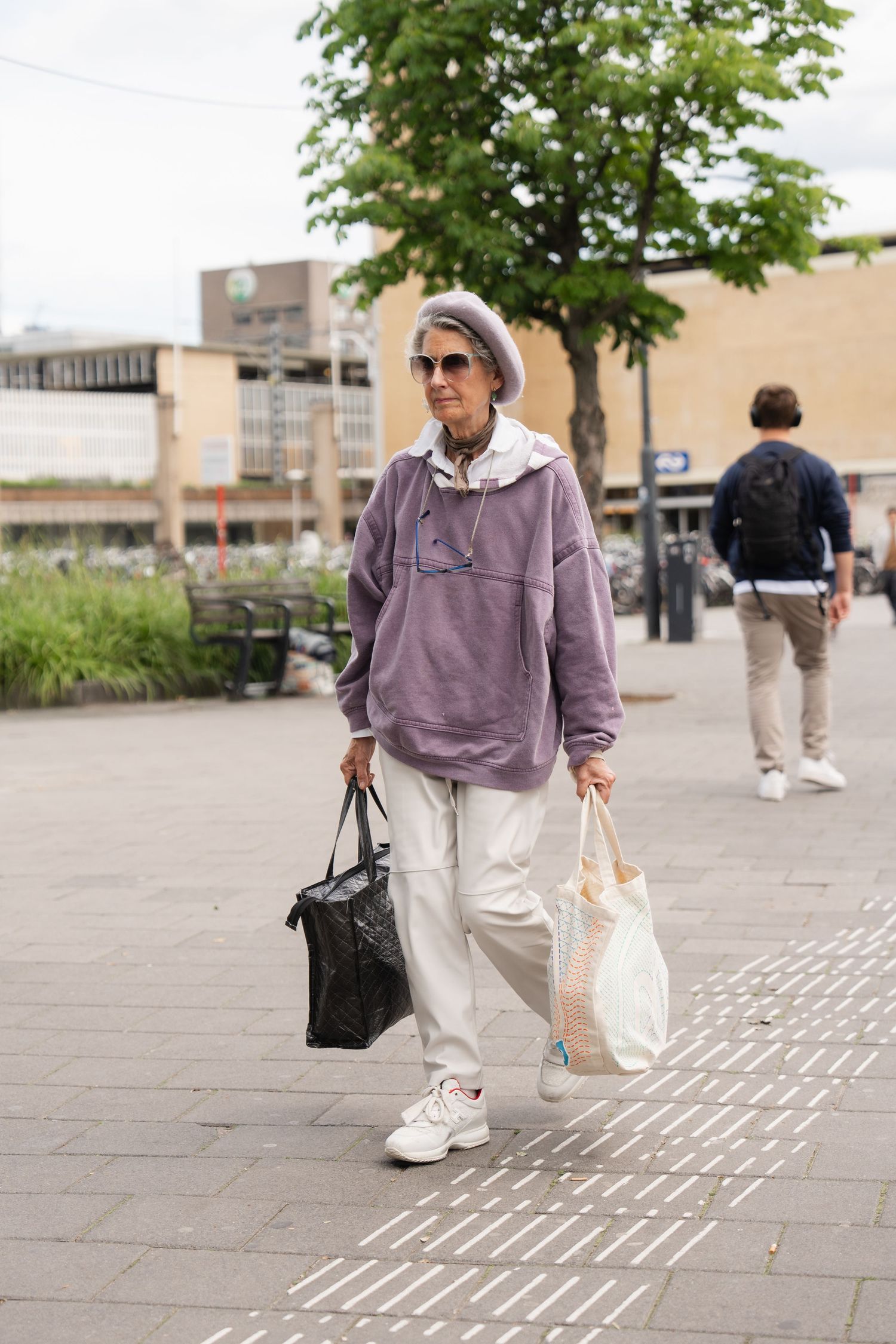Stylish senior woman in purple hoodie and white pants carrying bags while walking through an urban plaza.