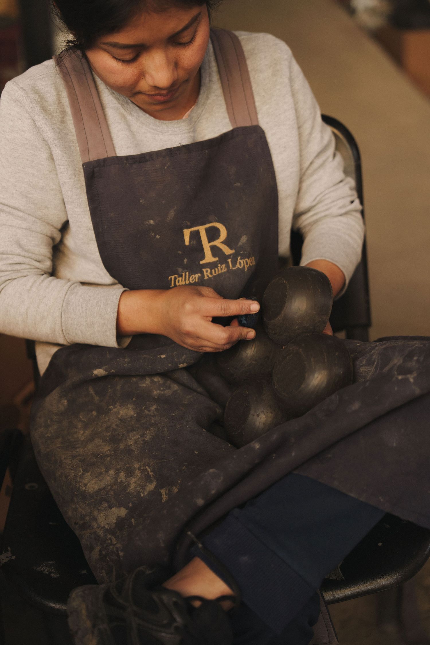 A craftswoman in a Taller Ruta López apron carefully shapes dark clay pottery pieces by hand in a workshop.