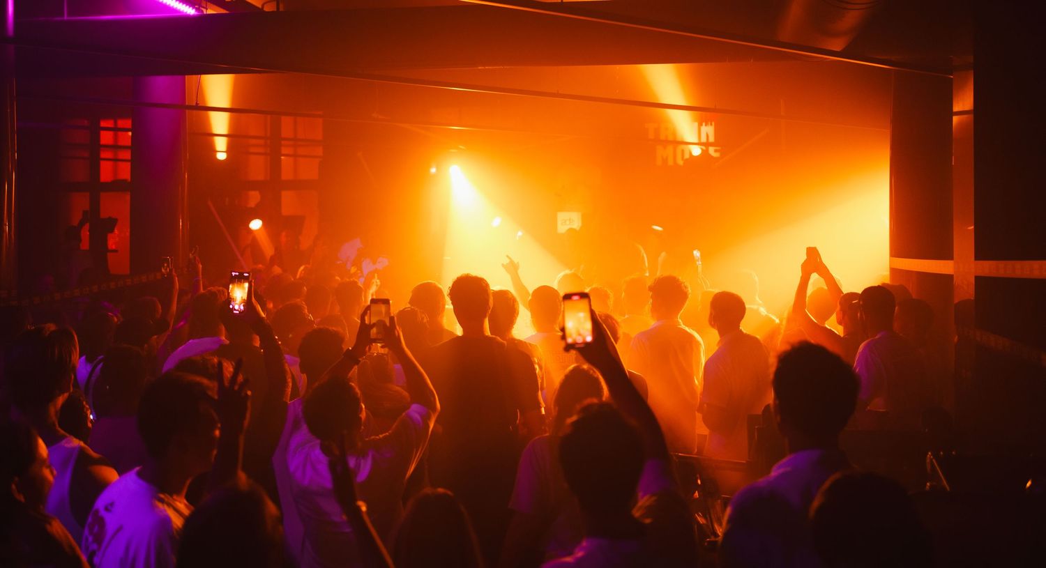 Crowded nightclub dance floor with orange stage lights and silhouetted partygoers holding up smartphones.