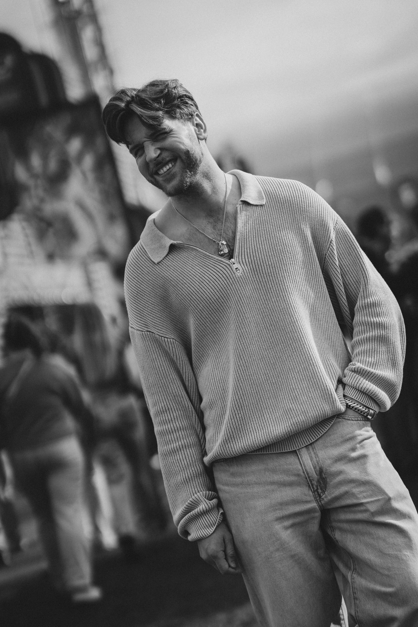 Black and white photo of a smiling man in a ribbed polo sweater and jeans, posing casually outdoors.