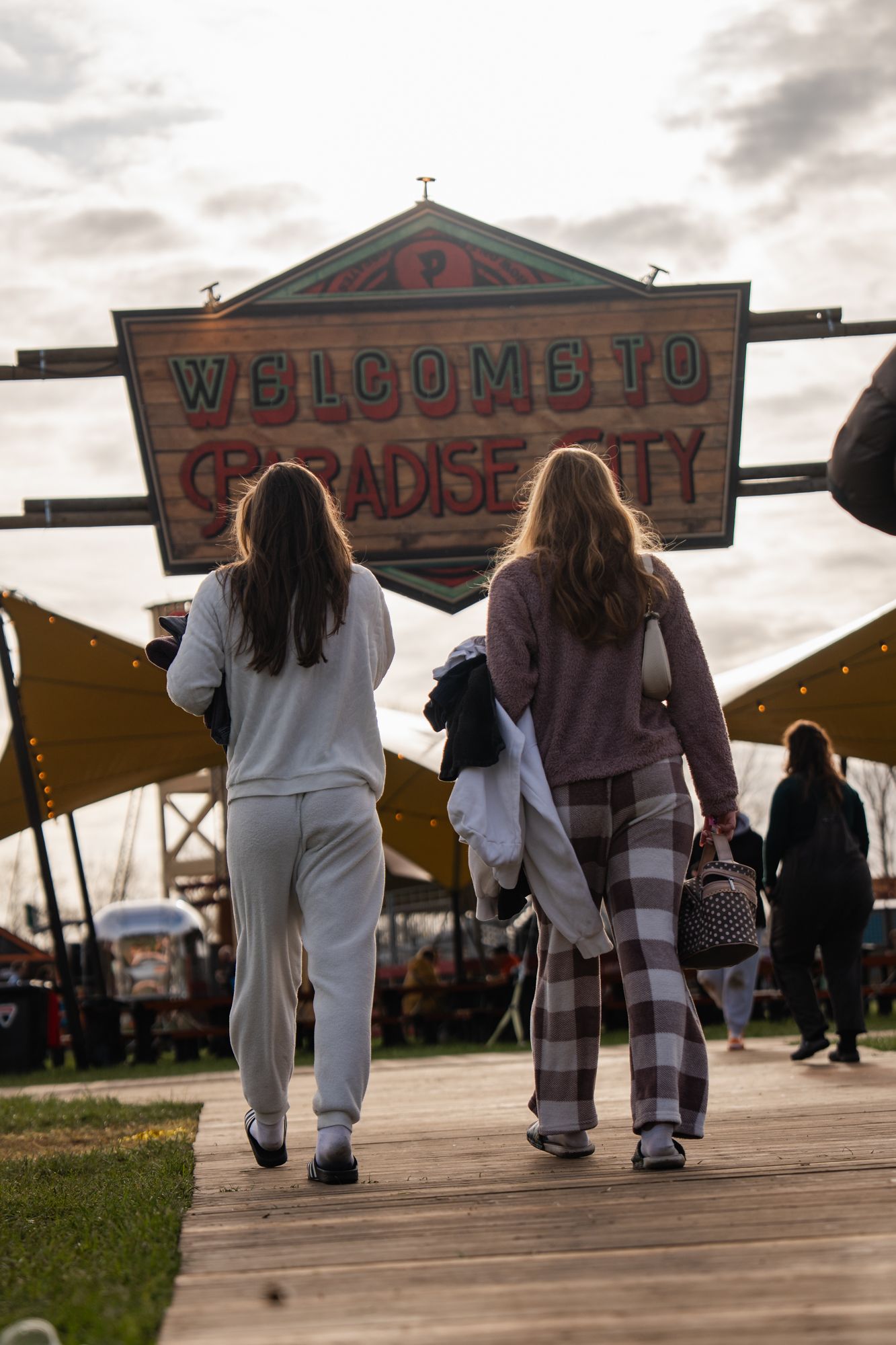 Two women walking toward a Welcome to Paradise City festival sign on a wooden boardwalk at dusk.