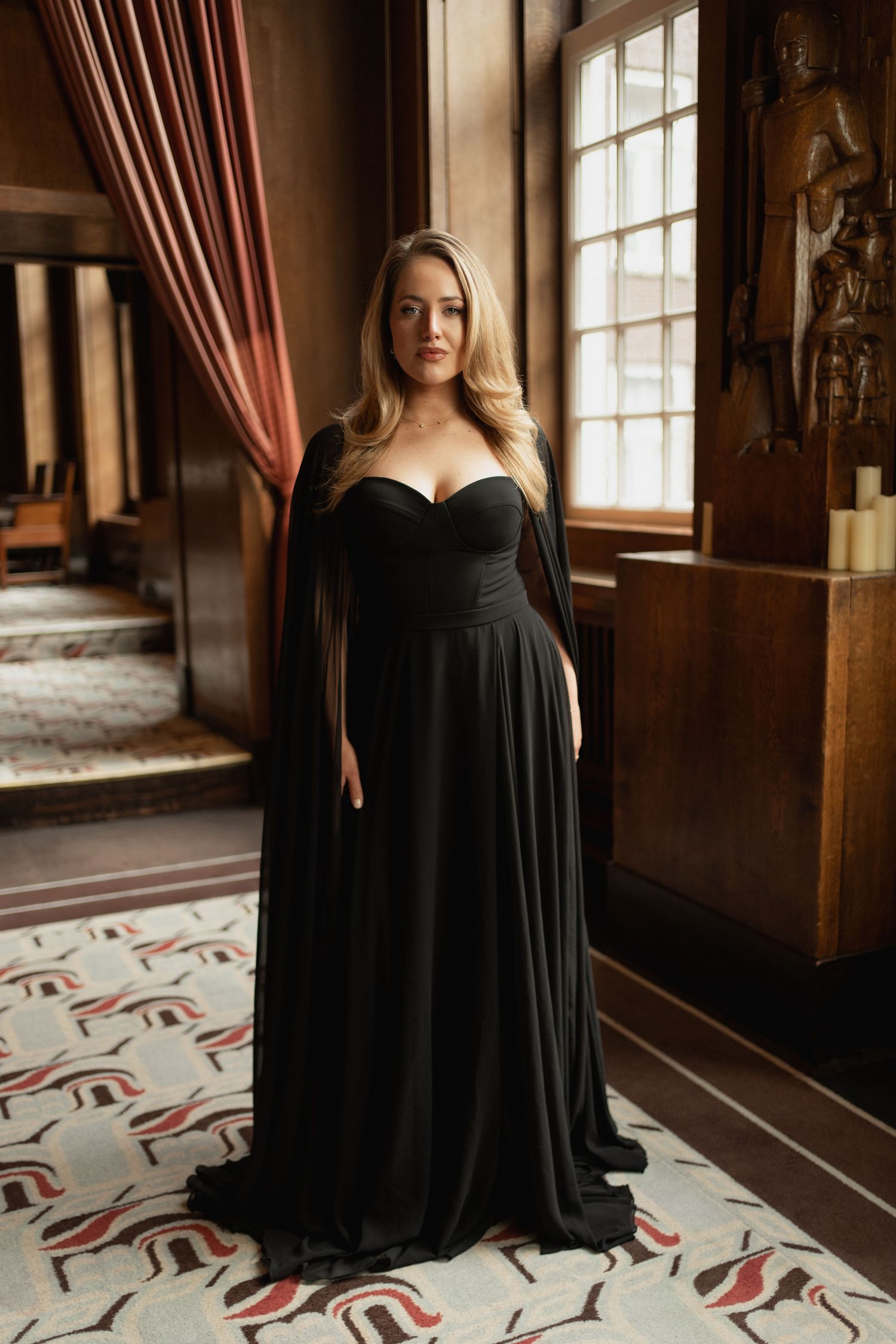 Woman in elegant black strapless gown with cape sleeves poses in ornate wood-paneled room with large window.