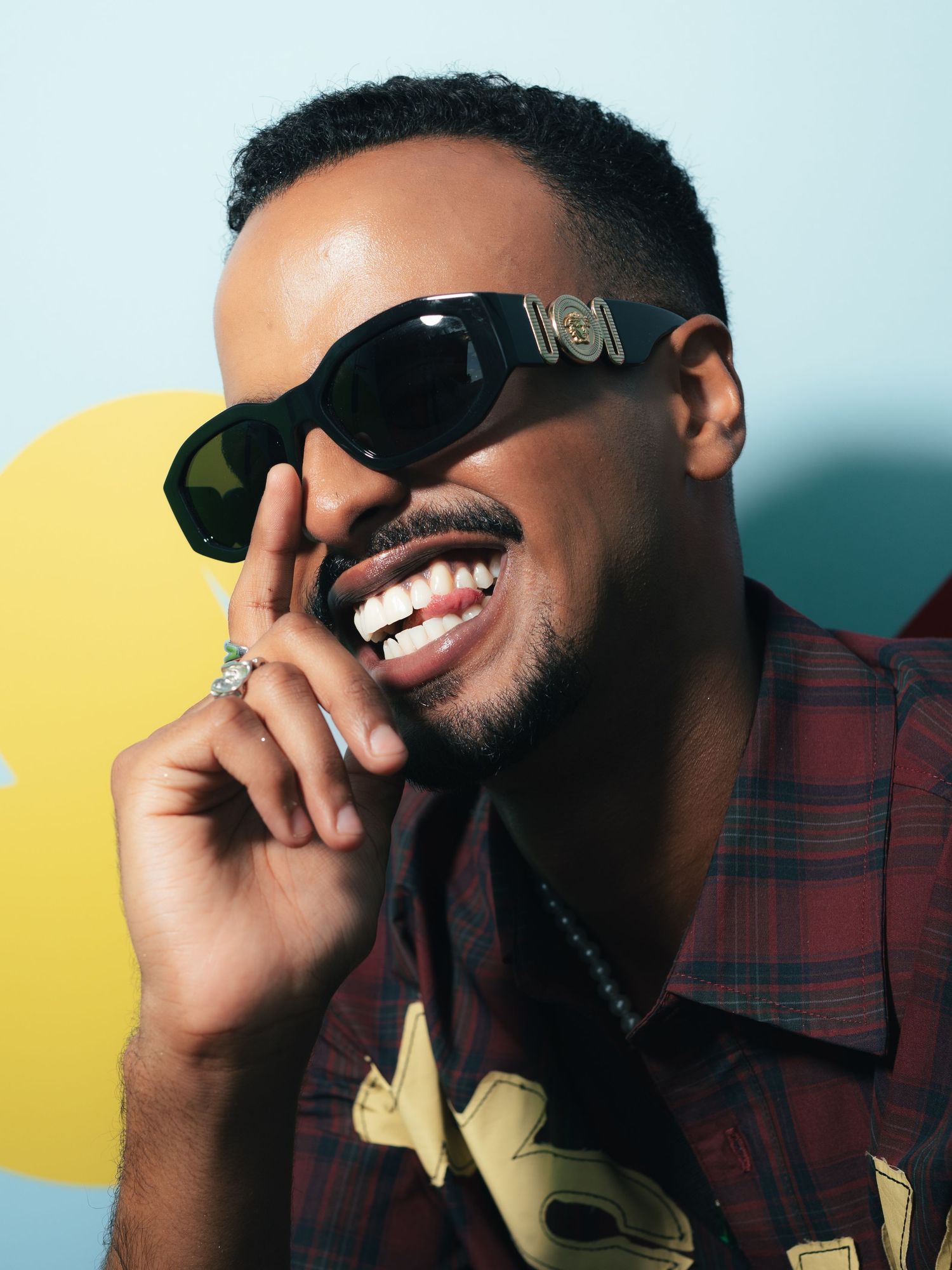 Smiling man wearing stylish dark Versace sunglasses and plaid shirt, laughing joyfully against a light blue background.
