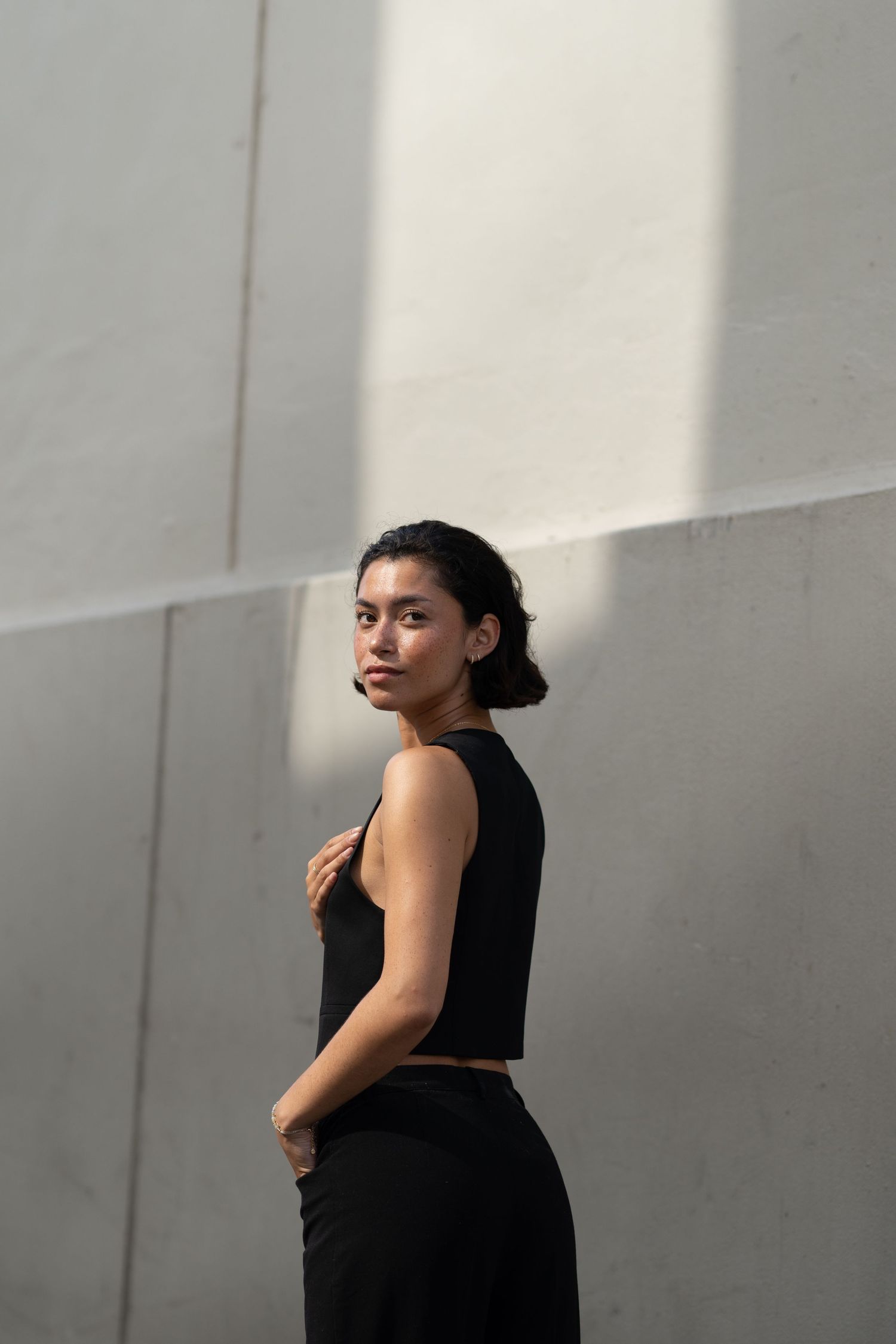 A young woman in a black crop top and pants poses confidently against a concrete wall in natural light.
