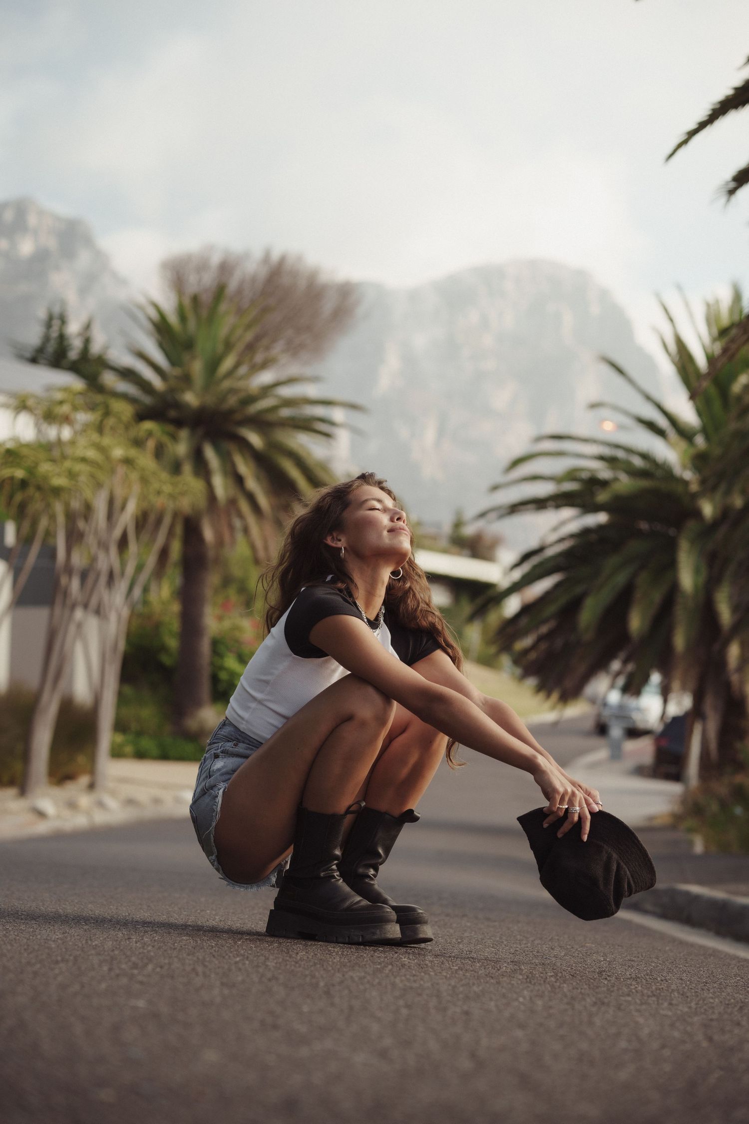 Young woman crouching on a road holding a hat, surrounded by palm trees and mountains in the background.