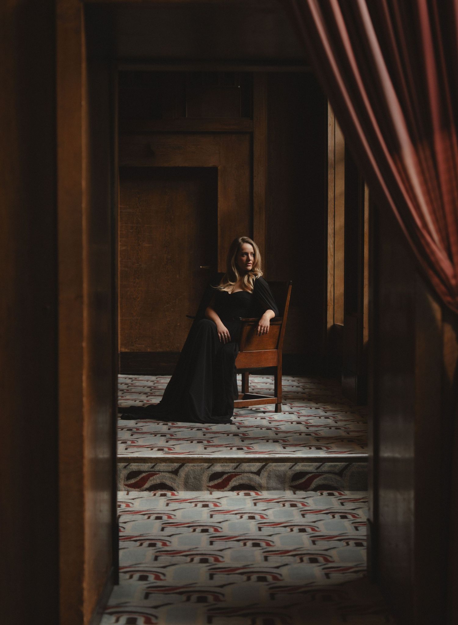 Woman in black gown seated on wooden chair in elegant room with patterned floor and red curtains.