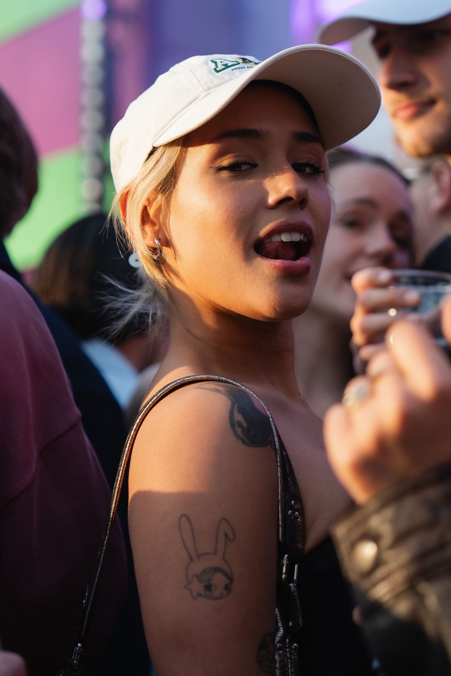 Young woman in white cap with bunny tattoo on shoulder smiling at outdoor event with crowd behind her.