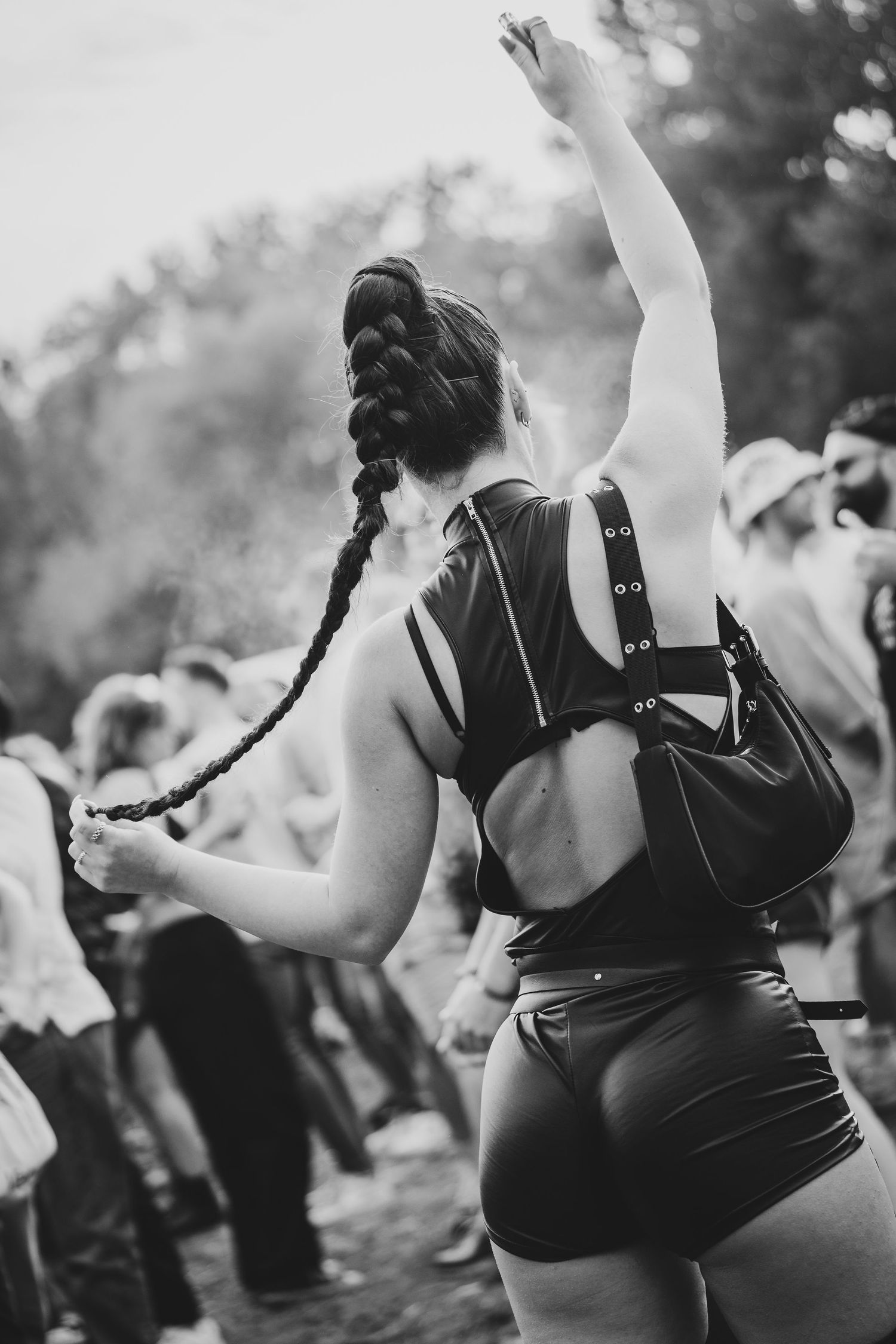 Black and white photo of a woman with braided hair dancing at an outdoor festival, wearing shorts and a backpack.
