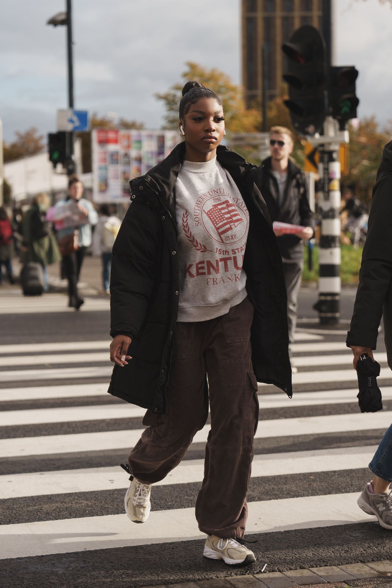 Woman in black puffer coat and Kentucky sweatshirt crossing a zebra crossing on a busy urban street.