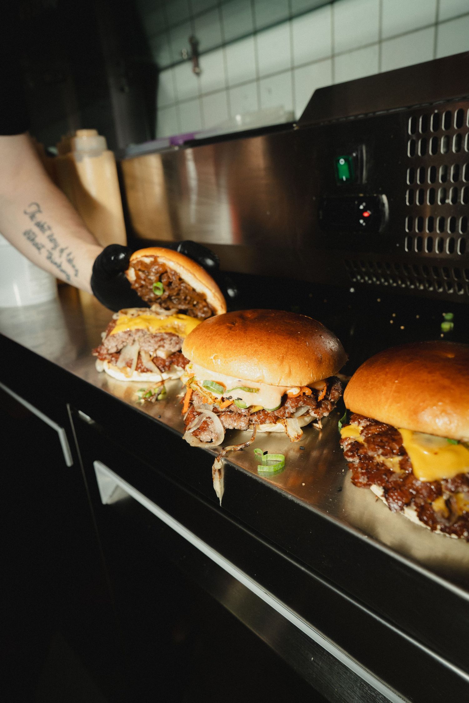Tattooed chef holding a juicy smash burger with melted cheese on a stainless steel kitchen counter.
